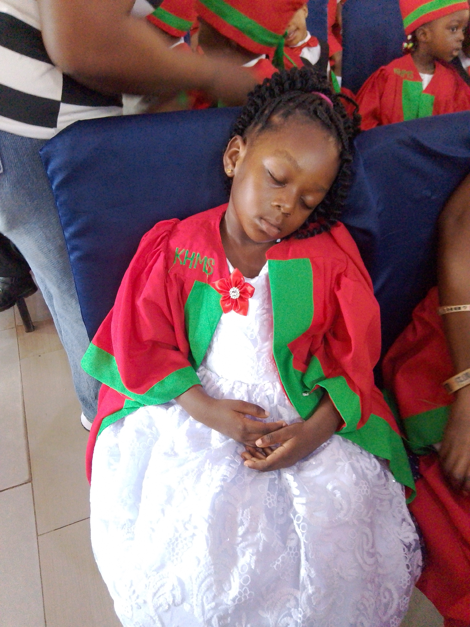 In Yenegoa, Nigeria, a young girl sleeps on a blue upholstered seat. She wears a white lace dress and a red robe with green trim, adorned with a red floral brooch. The robe has the letters "KHMS" embroidered in green on the left shoulder. Her hands are clasped in her lap. Behind her, other children in similar red and green robes can be seen. The floor appears to be tiled. The overall scene suggests an event or ceremony involving children.