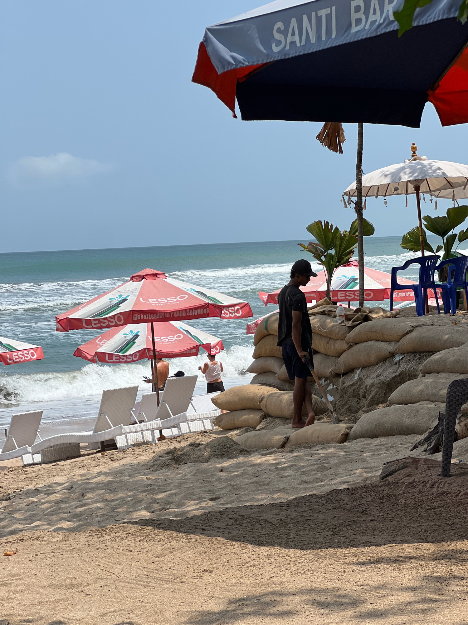 The image depicts a beach scene in Kuta, Indonesia. In the foreground, sand is piled up, reinforced with numerous sandbags, suggesting a barrier against the sea. A person wearing a dark t-shirt and shorts, with a cap on backwards, is standing on the sandbags, holding a shovel. Several white lounge chairs are arranged on the sand near the water's edge.
In the mid-ground, the ocean waves are crashing onto the shore. Several large, red and white umbrellas with the "LESSO" logo are positioned on the beach, providing shade. In the background, the sky is clear with a few clouds. There are also two blue chairs and more umbrellas, including a white one with tassels, and palm trees. The overall impression is that of a beach resort area, possibly preparing for or recovering from high tides.
