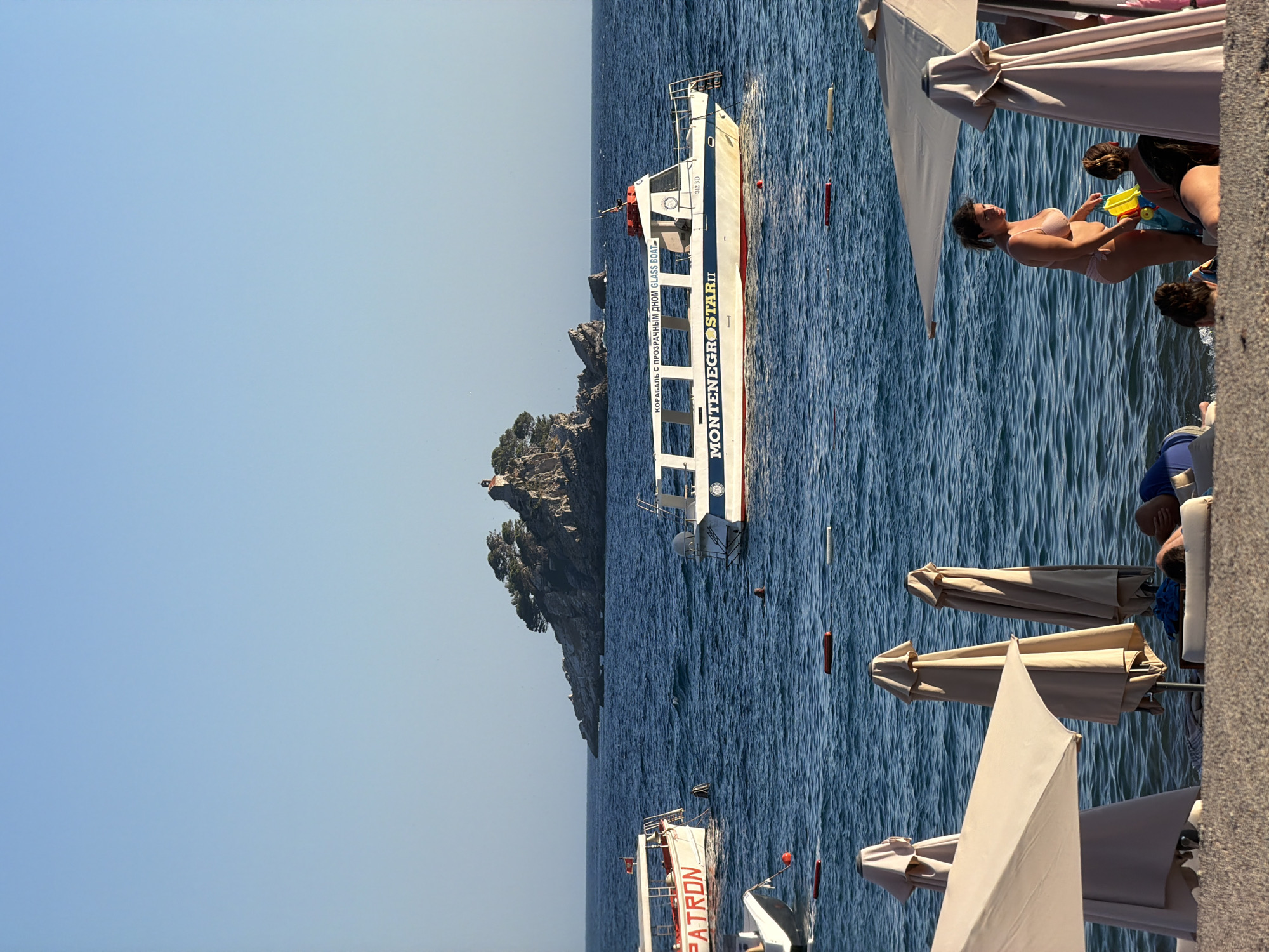 The image depicts a scene on the beach in Petrovac centar, Montenegro. In the foreground, beach umbrellas and lounge chairs are visible on the sand. Several people are present, including a woman in a pink bikini holding a toy, and other individuals relaxing on lounge chairs. In the middle ground, the blue sea is calm with gentle waves. Various boats are on the water, including a white boat with "MONTENEGRO STAR II" written on its side and another boat with "PATRON" on it. In the background, a rocky island with trees and a small structure on top is situated in the sea. The sky is clear and blue, indicating a sunny day.