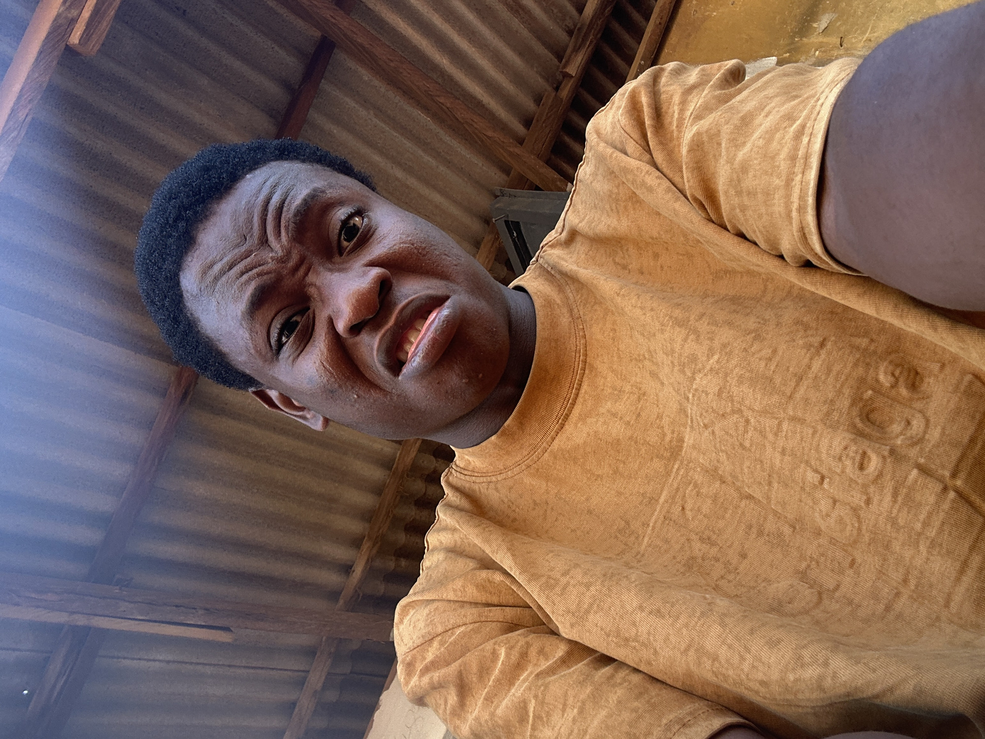 The image is a low-angle selfie featuring a young man with dark, short curly hair and brown eyes. He is looking directly into the camera with an exaggerated, humorous expression, characterized by a scrunched nose, deeply furrowed brow, and slightly open mouth revealing his teeth. Prominent wrinkles are visible across his forehead due to the intense facial expression.
He is wearing a mustard yellow or light brown t-shirt that appears to have a textured, possibly acid-washed or stonewashed finish. Upon closer inspection of the fabric, faint raised patterns and text are discernible, including "1980S", "57", "R", and other partially obscured letters or symbols.
The background shows the underside of a roof structure. It features parallel corrugated metal sheets supported by exposed wooden beams and rafters. This utilitarian setting suggests the photo was taken indoors or under a simple covered outdoor area. The lighting indicates it is daytime. The man's expression conveys a sense of playful humor or mock-discomfort.