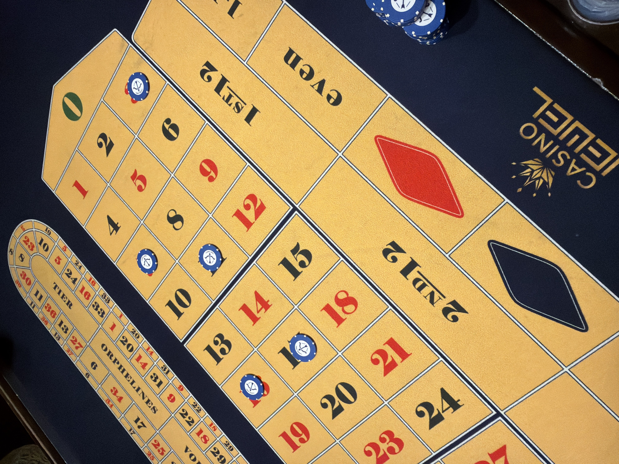 The image shows a roulette table at Casino Jewel in Tbilisi, Georgia. The table is covered with a yellow felt mat featuring a grid of numbers and betting areas. Several blue casino chips are visible, with one placed on the number '2' and another on the number '10'. The "0" is green, and the numbers 1 through 36 are in red and black. The betting areas include "1st 12", "2nd 12", and "even". The "Casino Jewel" logo is visible in the bottom right corner. The lighting appears to be artificial, likely from overhead casino lights, casting shadows on the felt. The perspective is from a high angle, looking down at the table. No people are visible in the image.