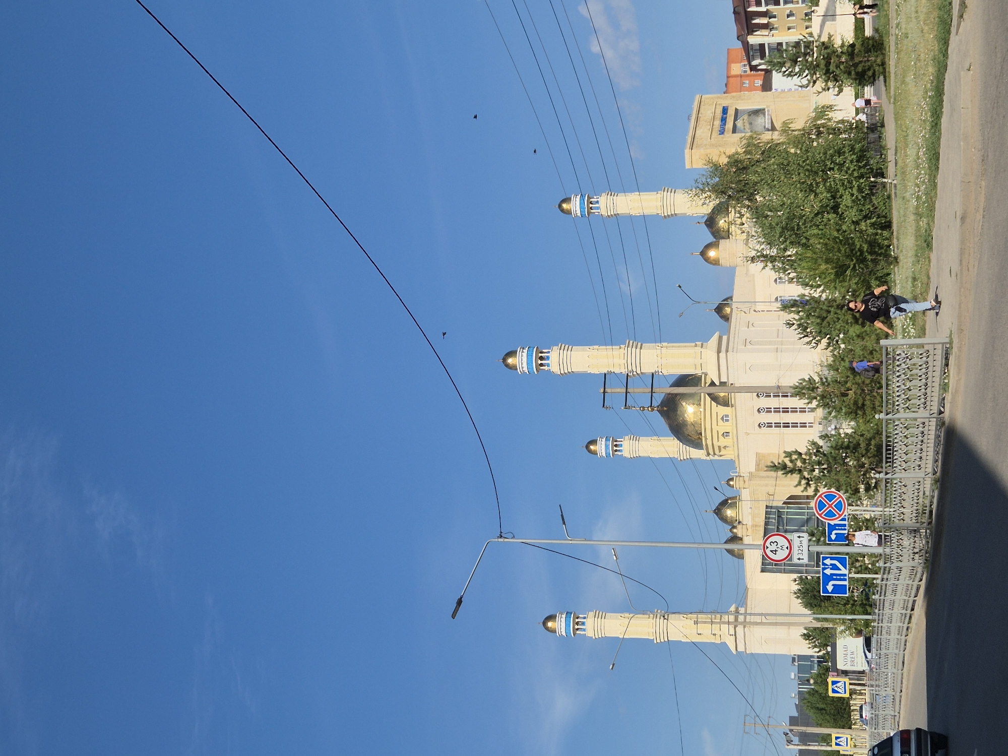 The image captures a sunny day scene in Kokshetau, Kazakhstan, featuring the large "Kokshetau Central Mosque" as the prominent subject. The mosque is a light-colored building with multiple golden domes and tall, slender minarets topped with blue accents. In the foreground, a paved road runs horizontally across the lower part of the frame, with a pedestrian walkway and a decorative metal fence separating it from a grassy area.
Several street signs are visible along the walkway, including directional arrows and a speed limit sign indicating "43 M" and "325M". A woman wearing a black t-shirt and blue jeans is walking on the walkway, looking towards the left. Behind her, a few other individuals are walking and standing. Lush green trees and bushes are interspersed throughout the scene, adding to the natural beauty. In the background, residential buildings and other structures suggest an urban environment. The sky is a clear, vibrant blue, with a few wispy clouds and power lines crisscrossing the upper portion of the image. The overall atmosphere is serene and peaceful, indicative of a typical day in the city.