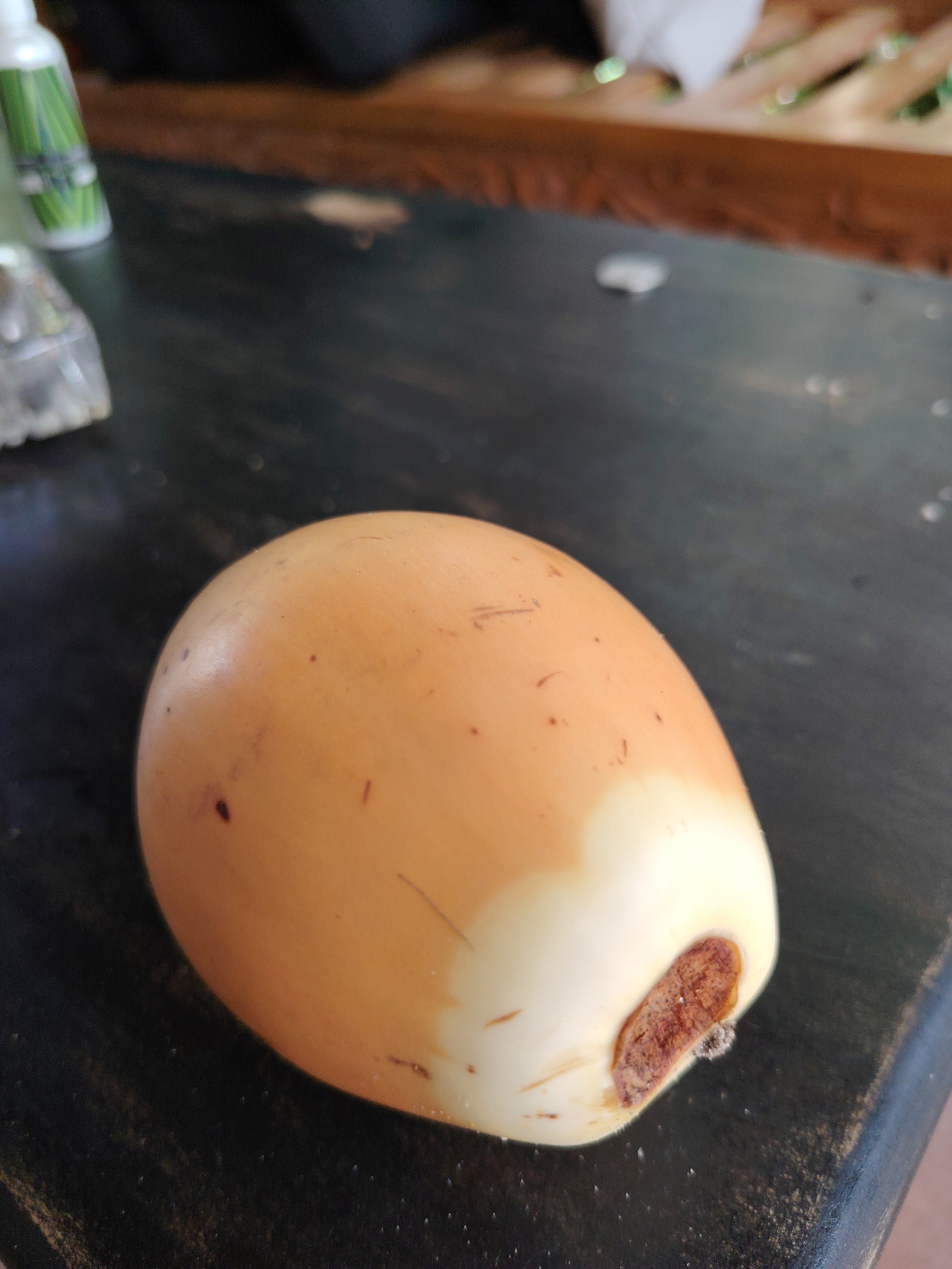 The image displays a close-up of a single, young, pale orange-yellow coconut resting on a dark, speckled surface. The coconut is smooth, with minor dark specks and faint linear markings across its surface. One end of the coconut is distinctly lighter, almost white, with a prominent, rough, dark brown, oval-shaped attachment point. A tiny speck of white, possibly mold or fluff, adheres near this brown area. The dark surface beneath the coconut appears dusty or textured with small, light-colored particles.
In the blurred background to the left, a white bottle with an unidentifiable green label and a clear plastic item are partially visible. Further back and to the right, a brown, slatted structure, possibly wood, is out of focus. The overall lighting suggests natural daylight. No human subjects or discernible activities are depicted. The scene conveys a casual, possibly outdoor or semi-outdoor setting. No legible text is visible.