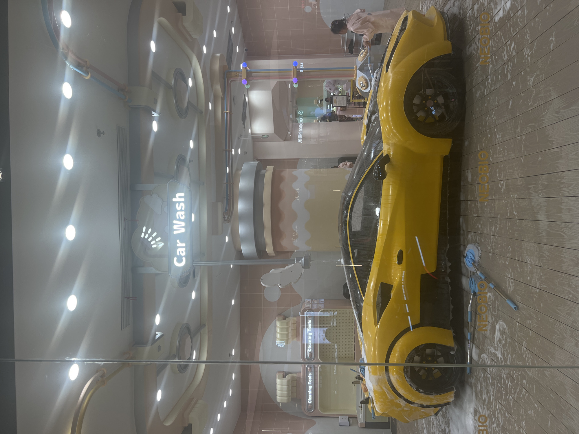 The image shows a yellow sports car with a black roof, parked on a wet, wooden floor covered in soap suds, likely at a car wash. Two mops are lying on the floor near the car's rear wheel. In the background, there are two stations labeled "Cleaning Tools." Above the car, a sign reads "Car Wash." The scene appears to be indoors, with overhead lighting. A young girl in a pink robe is standing to the right of the car, facing away from the viewer. Other people are visible in the background, appearing to be children and adults, some of whom seem to be interacting with the car or equipment. The overall impression is that of a children's play area designed to simulate a car wash. The location is identified as Liangjiang, China.