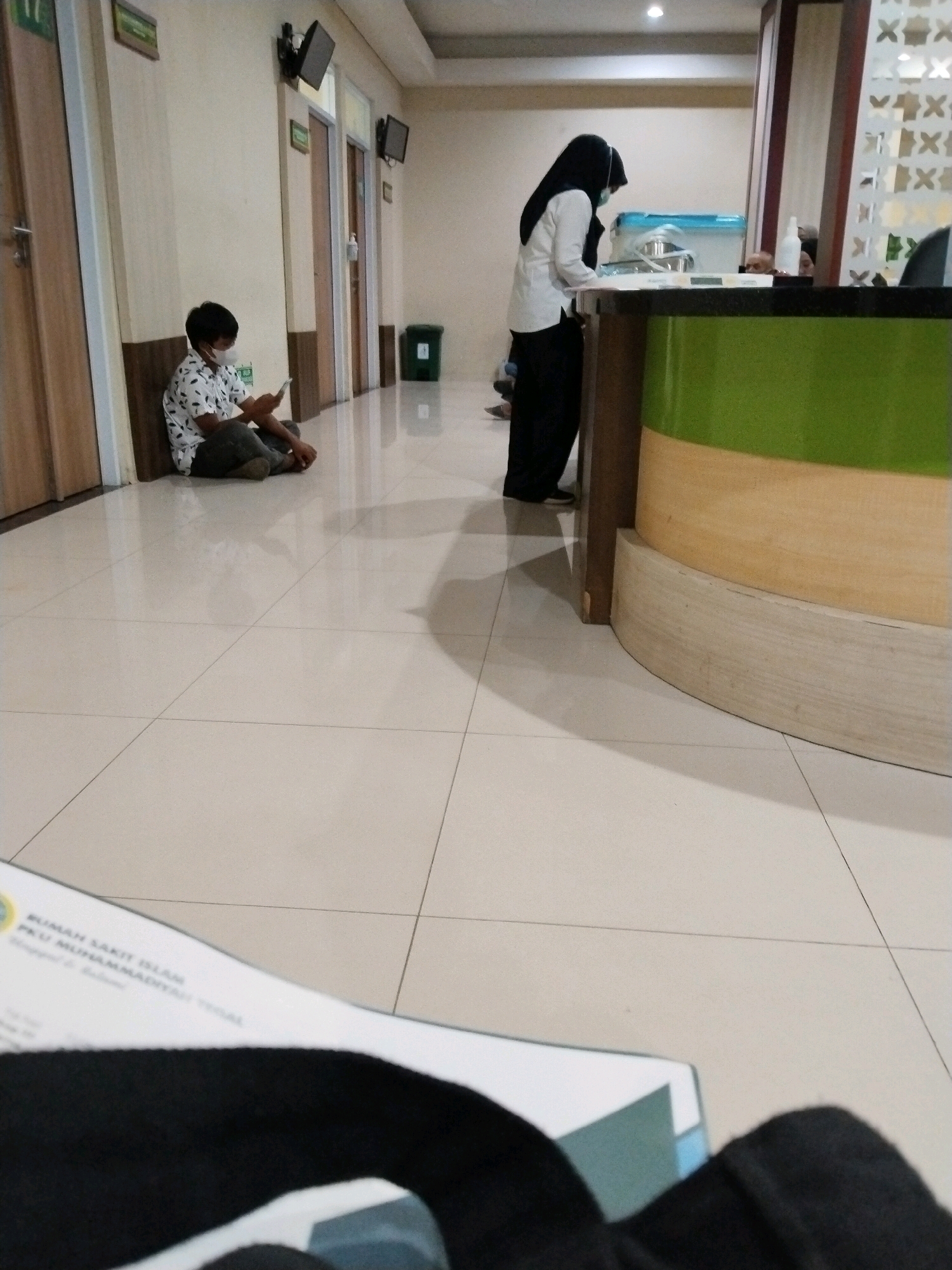 The image depicts a waiting area, likely a clinic or hospital, in Adiwerna, Indonesia. In the foreground, a blurred document with text is visible, suggesting it belongs to a patient or visitor. The main subject in the mid-ground is a young male, wearing a patterned short-sleeved shirt, dark pants, and a face mask, sitting on the floor with his back against a wall. He is holding a smartphone and appears to be engaged with it. To the right, a female individual in a black hijab and white top, also wearing a face mask, stands behind a counter. She is attending to something on the counter. The environment includes a tiled floor, a long counter with green and beige sections, wooden doors, and what appear to be monitors mounted on the wall. A green trash can is visible near one of the doors. The lighting suggests an indoor setting, possibly with artificial illumination. The overall scene conveys a sense of waiting and routine activity within a healthcare facility.