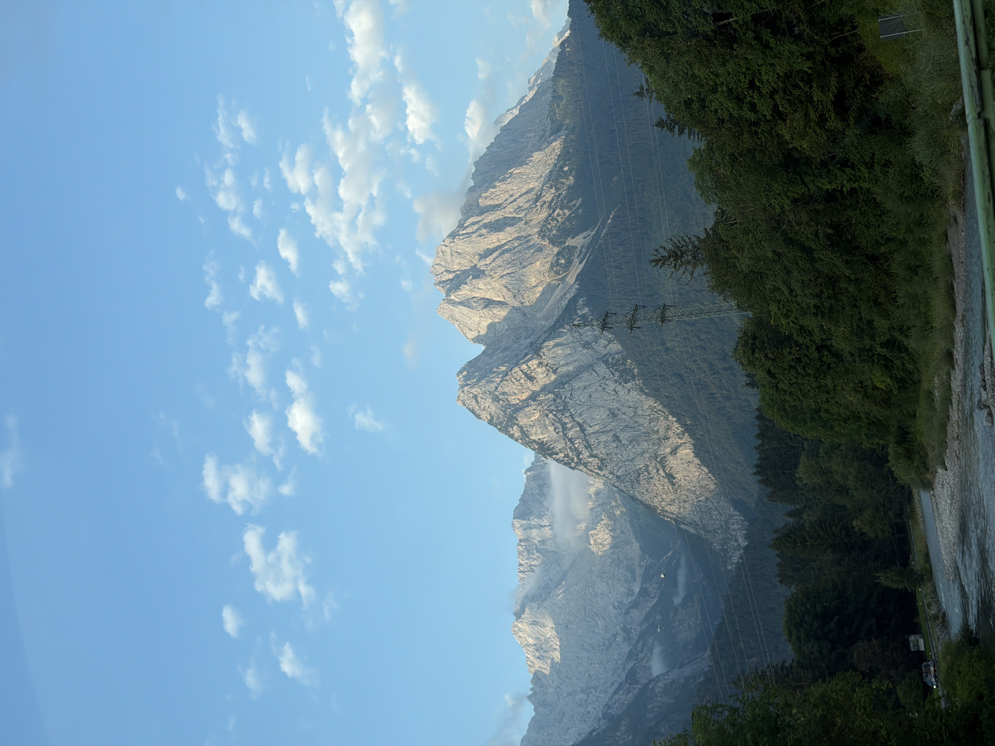 The image captures a panoramic view of the Bavarian Alps, likely near Garmisch-Partenkirchen, Germany, under a partly cloudy blue sky. In the foreground, a fast-flowing river with a gravelly bed winds through a verdant valley. The riverbanks are lined with lush, dark green trees, some of which are deciduous, indicating a summer or early autumn setting. A prominent, steep mountain range dominates the background. These mountains are characterized by their jagged, rocky peaks, with some areas illuminated by sunlight, highlighting their texture and scale. Patches of mist or low-lying clouds cling to the sides of some mountains, adding depth and atmosphere. In the midground, an electrical pylon with multiple wires stretches across the landscape, contrasting with the natural scenery. A narrow road or path is visible on the left side of the river, with a few vehicles present. The overall impression is one of a majestic natural landscape, with a hint of human infrastructure.