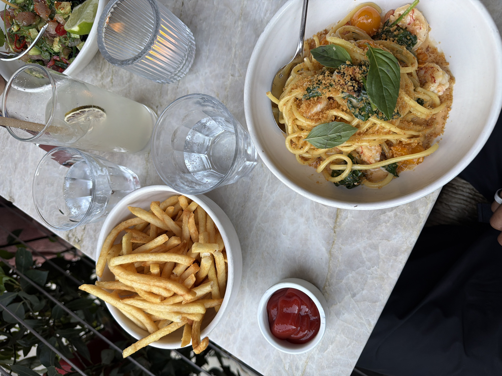 The image presents a bird's-eye view of a table laden with food and drinks, likely in a restaurant setting in West Hollywood, USA. A bowl of pasta with shrimp, tomatoes, and basil sits at the upper right. A side of french fries in a white bowl is at the bottom left with a small dish of ketchup to its right. A glass of lemonade sits center left along with two empty clear glasses and a votive candle holder. At the upper left, there is a bowl of salad. The table has a marble-like top. A small section of a person's black shirt is visible in the lower right corner.