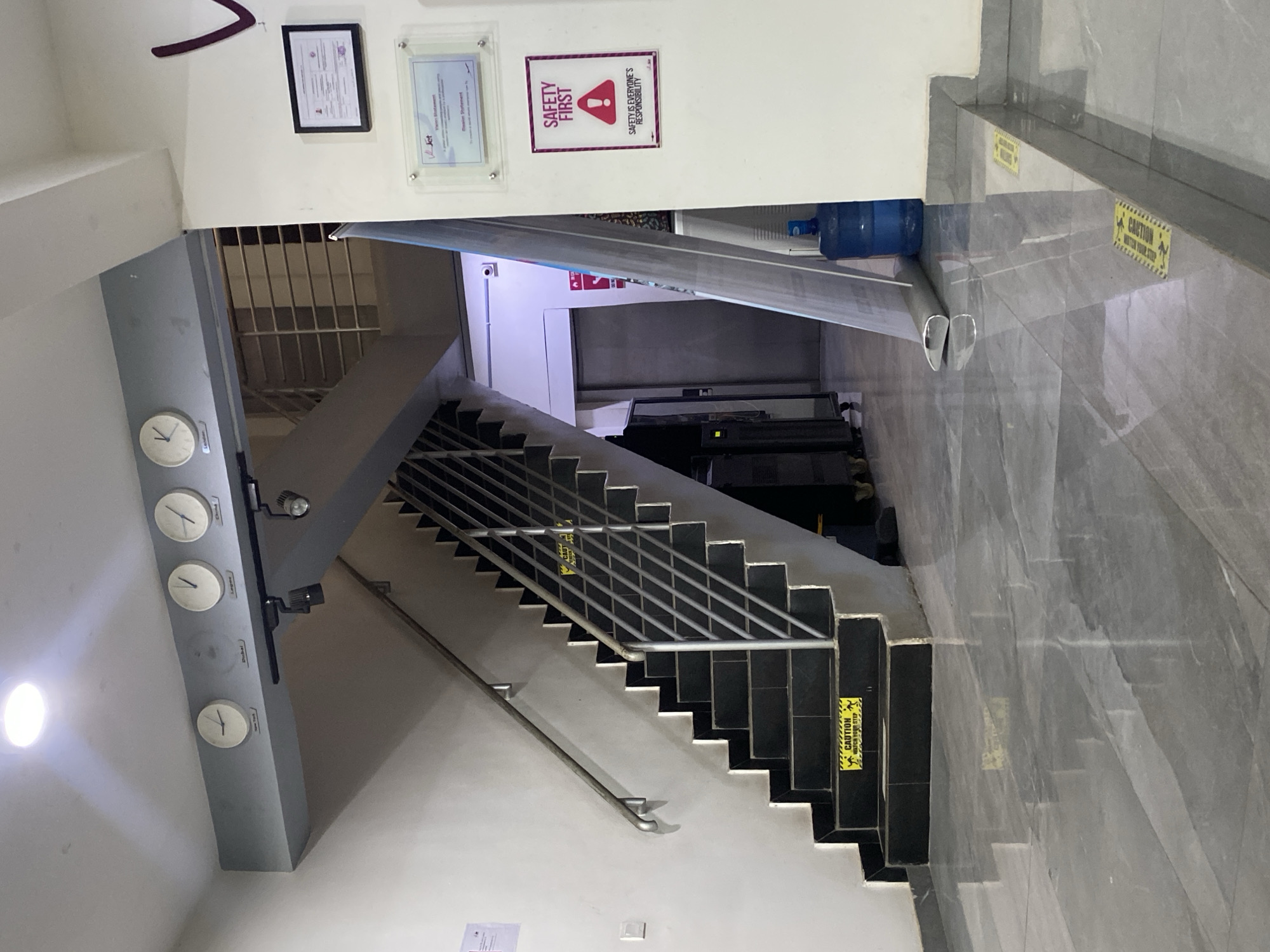 The image shows an interior view of a building, likely in Ikeja, Nigeria, given the presence of multiple clocks displaying different international times, including Lagos (Nigeria). The scene is dominated by a staircase with black treads and white risers, leading downwards. A metal railing runs alongside the stairs. Wall-mounted clocks, four in total, are present.
Several signs are visible on the walls. Near the top of the frame, two framed documents and a "Safety First" sign are mounted on the wall. "Caution: Watch Your Step" warning labels are placed on the steps and on the floor near the top of the stairs.
At the top of the stairs, a blue water cooler bottle is visible. The floor is tiled in grey marble pattern. The lighting appears to be artificial. The image is taken from an elevated position, looking down the stairs.