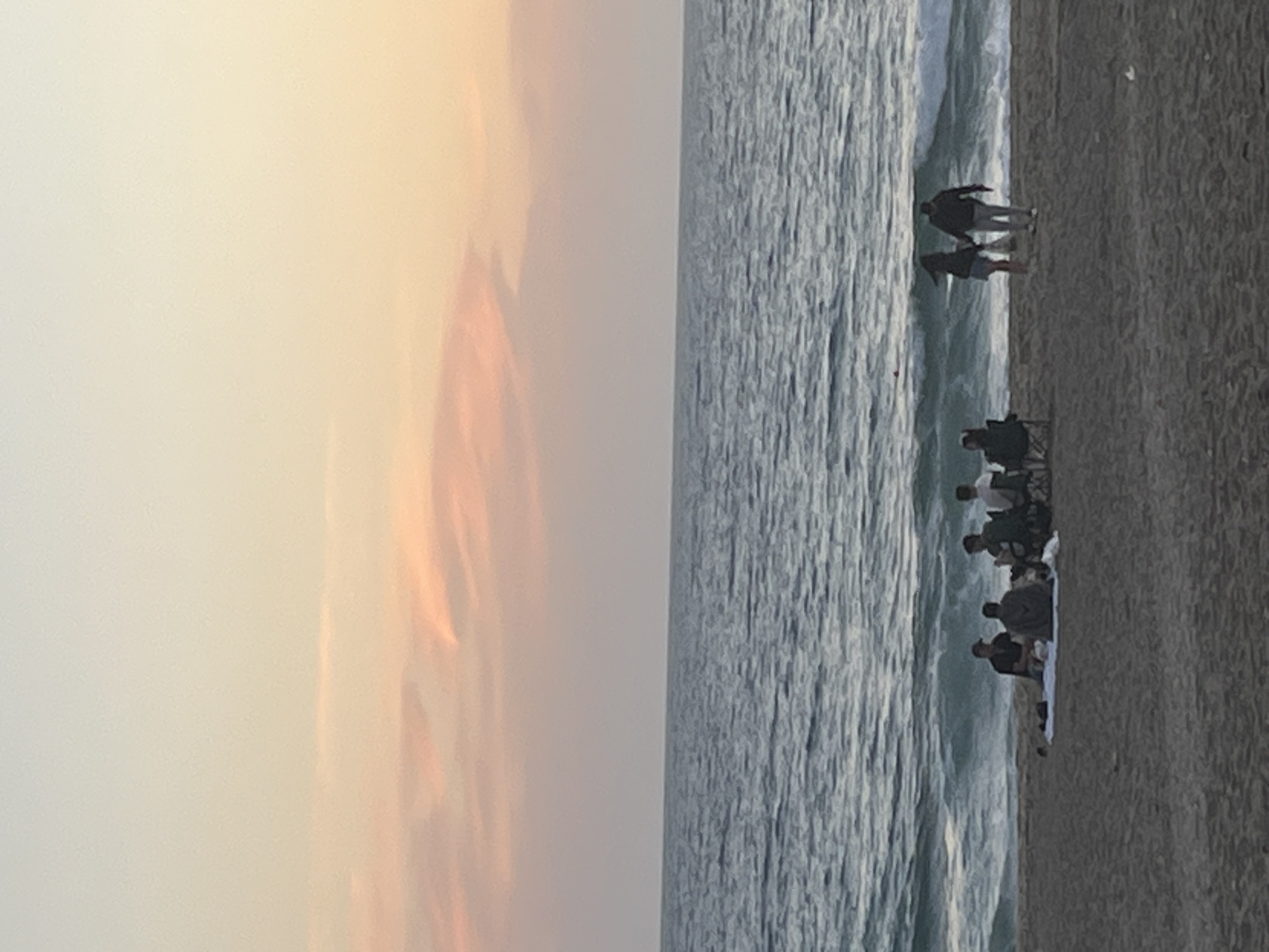 The image shows a beach scene at dusk, likely in Alanya, Turkey. The sky is a gradient of pale yellow to pink. The sea is moderately rough, with white-capped waves. On the beach, there is a group of people sitting and standing. Two people are standing at the edge of the water, and a group of about six people are sitting on the sand further up the beach, possibly on a blanket or towels. The sand is a dark beige color. There may be a chair among the group of people.