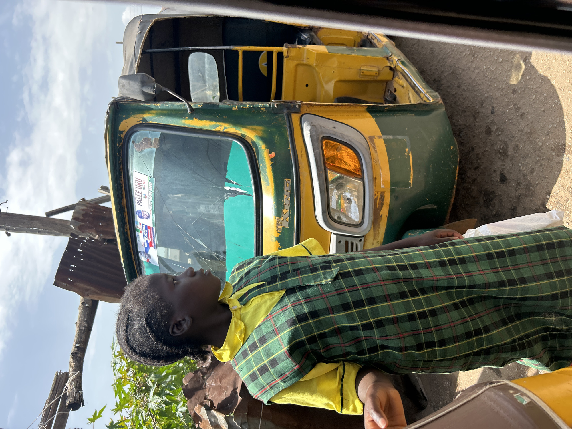 A young Black person, dressed in a yellow collared shirt and a green and black plaid vest, stands near the front of a weathered yellow and green auto-rickshaw. The auto-rickshaw appears to be parked on a dirt and gravel road, with some scattered debris, including corrugated metal, visible around it. The person is looking upwards and to the left with a neutral expression, their dark hair styled in braids. The auto-rickshaw has some visible wear and tear, with rust and faded paint. The windshield has a sticker affixed to it, with visible text that appears to be in English and possibly another language, including names that might be political or campaign related, and a portrait of a person. The sky above is bright and cloudy, suggesting it is daytime. The overall scene suggests an outdoor setting in a somewhat rural or informal urban area. The time of day appears to be daytime due to the lighting and shadows. The location cues, such as the type of vehicle and the debris, are consistent with a setting in Jos, Nigeria.