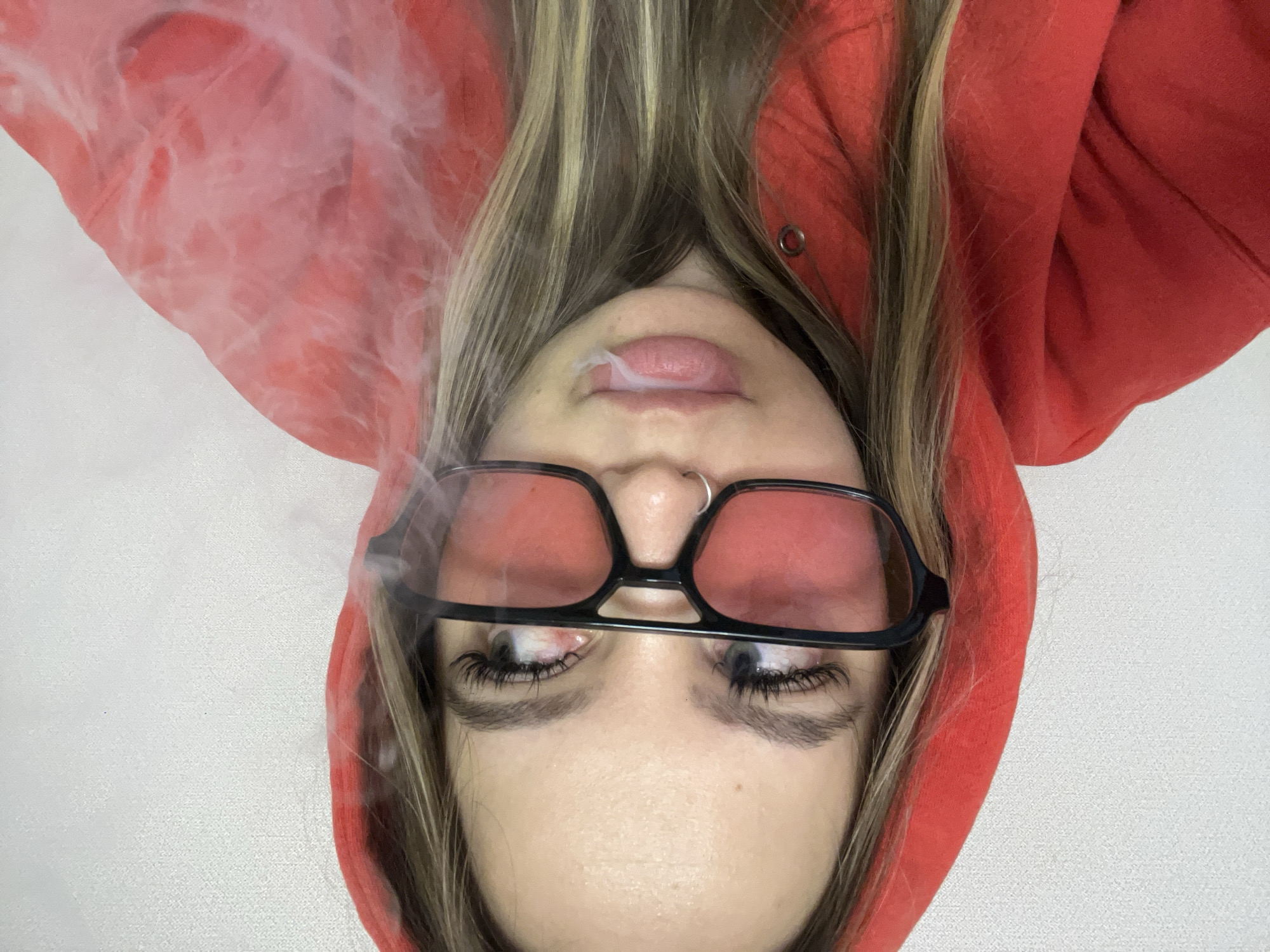A woman with long blonde-streaked hair, wearing black-rimmed glasses with a red tint and a red hoodie, is lying on her back against a white surface, exhaling smoke from her mouth. She has a nose ring. The location is not discernible.