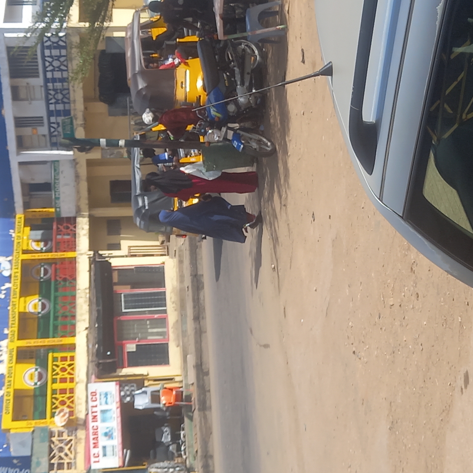 This media file depicts a busy street scene in Dadi Riba, Nigeria, under bright daylight, evidenced by pronounced shadows. The setting features an unpaved, dusty road lined with multi-story buildings of varying architecture.
Several individuals are visible, including two women walking on the street in traditional attire; one wears a red dress with a black hijab, and the other is in a blue garment and hijab. A black motorcycle is parked on the side. The scene includes multiple yellow Keke Napeps (motorized tricycles), some appearing to be parked, and one with people near it. Part of a silver car is visible on the right, featuring a dark tinted window and an extended antenna.
Buildings display diverse features, including a structure on the left adorned with yellow and green panels, circular motifs, and red crisscross patterns. Visible text from signs and banners includes "OFFICE OF YAN DOYA CHAPEL ROAD TRANSPORT EMPLOYER'S ASSOCIATION OF NIGERIA," "LC. MARC INT'L CO.," "DE MODELOVA," and "OPDANF." Arabic script "الري" is also present. A street pole shows the number "40." The image portrays an active, typical urban or semi-urban environment.