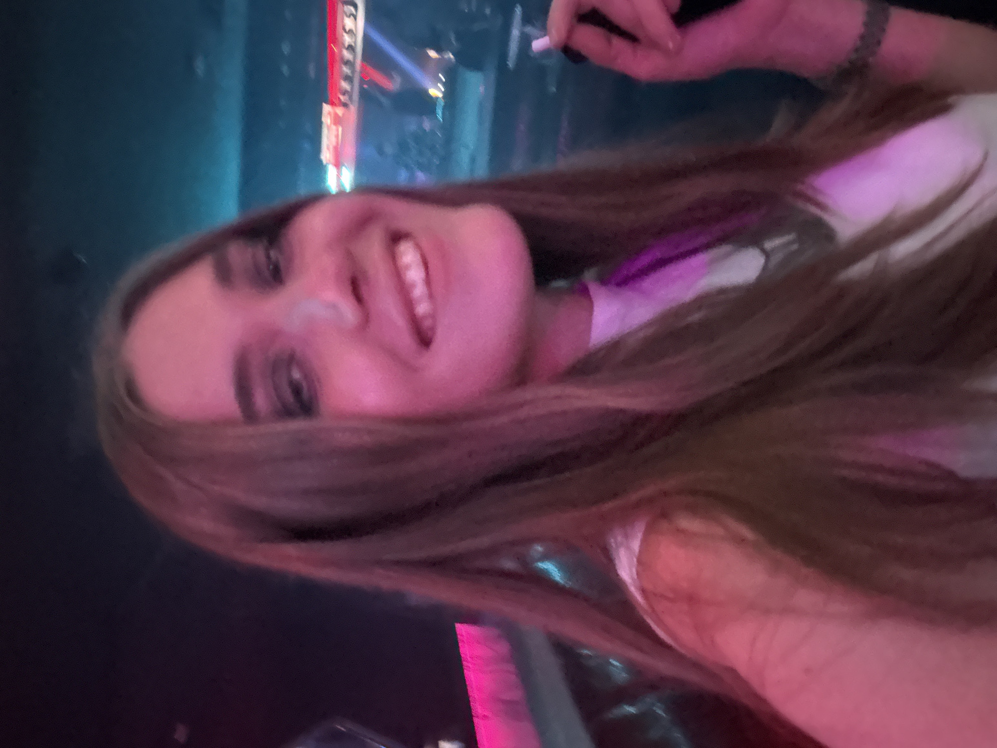 The image displays a close-up, chest-up portrait of a young adult female situated within a dimly lit entertainment venue in Mexico City, Mexico. The subject, facing forward and slightly to the left, has long, light brown hair extending over her shoulders. She is smiling broadly, exposing her teeth, and wears dark eye makeup. She is dressed in a white top. Her right hand, positioned in the lower-left quadrant of the frame, holds a small, white cylindrical object, consistent with a vape pen or cigarette, between her index and middle fingers; her fingernails are painted dark. Her left wrist, partially visible, bears a dark bracelet or tattoo. The background is significantly out of focus, characterized by various artificial colored lights including prominent pink/magenta illumination on the left and blue/cyan lighting on the right, with additional distant red light sources, indicative of a nightclub or concert environment.