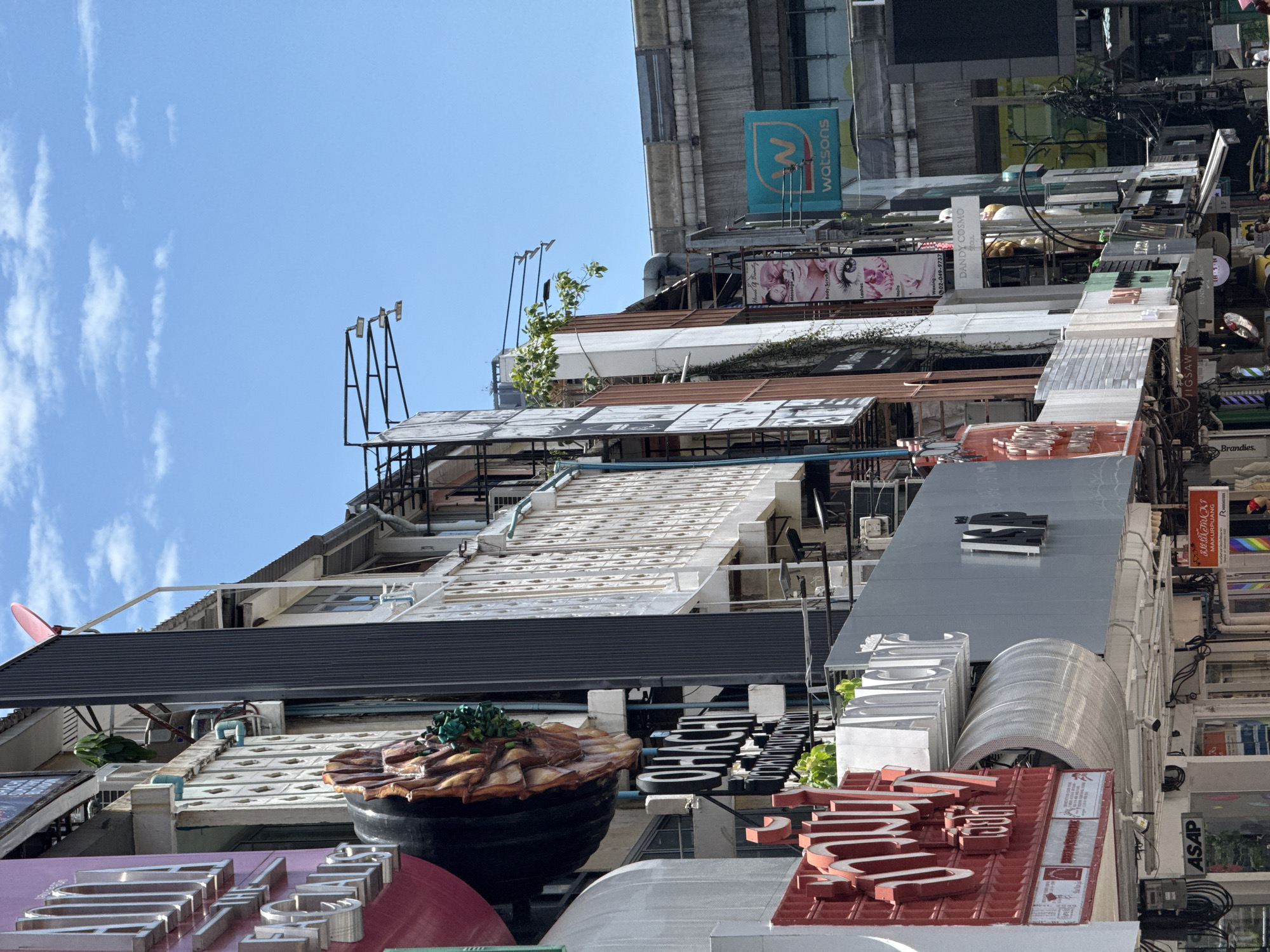 The image is a street-level view looking up at the facades of several buildings in Bangkok, Thailand. The architecture is a mix of modern and older structures, with prominent signage and awnings. In the foreground, a red sign with white lettering is visible on the left. To the right of this, a silver sign that reads "OHACHI" is mounted on a dark background. Further to the right, a teal sign with a "W" logo and the word "watsons" indicates a retail establishment. Buildings are multi-story, with balconies and exposed utility wires present. The sky above is clear blue with scattered white clouds. The scene depicts a busy commercial street.