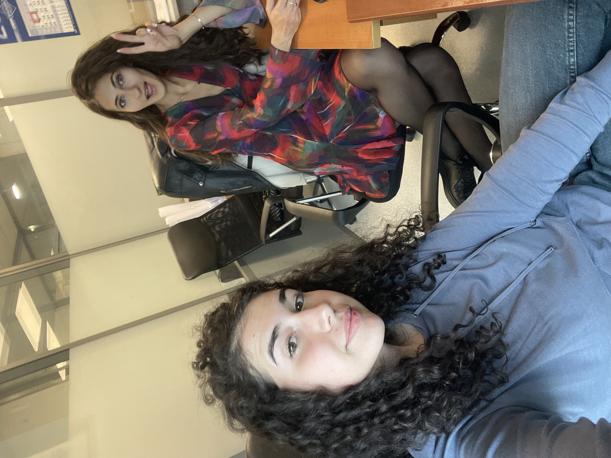 The image, rotated for proper viewing, depicts two young women in a modern office environment in Sofia, Bulgaria, possibly posing for a casual photo or a selfie.
In the foreground, on the right side of the frame, a woman with dark, curly hair and a light blue hoodie is visible. She wears blue jeans and looks directly at the camera with a slight, friendly smile.
Behind a wooden desk, positioned on the left side of the frame, sits another woman with long, dark, wavy hair. She is dressed in a vibrant, multi-colored long-sleeved dress featuring patterns of red, purple, blue, and green. She also wears sheer black tights and black shoes. Her right hand is raised, forming a peace sign, and she displays a wide, cheerful smile, looking towards the camera. A ring is visible on her finger and a bracelet on her wrist. A black leather jacket is draped over a black office chair near her.
The background reveals typical office elements, including light-colored walls and glass partitions that reflect interior lighting. A document or calendar with some faint, illegible text and numbers is attached to the wall behind the seated woman. Both women convey a relaxed and happy demeanor, contributing to a friendly atmosphere in what appears to be a daytime setting.