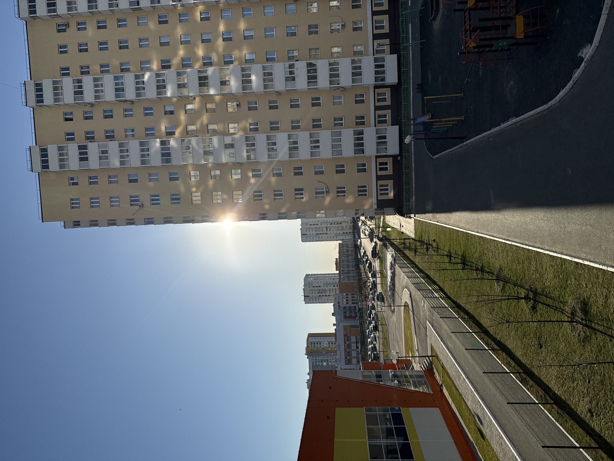 The image shows a view of an urban landscape in Tobolsk, Russia. A tall, multi-story apartment building with numerous windows dominates the left side of the frame, its facade reflecting the sunlight. Adjacent to this building is a children's playground with various play structures on a dark surface. A paved road stretches along the right side, bordered by a strip of green grass and young trees. In the distance, more buildings are visible, indicating a dense urban area. The sky is clear and blue, with the sun positioned behind the apartment building, creating a bright glare. A building with orange and yellow panels is visible at the bottom of the image. Parked cars can be seen along the road in the distance. The image appears to be taken from a high vantage point, possibly from another building or an elevated structure.