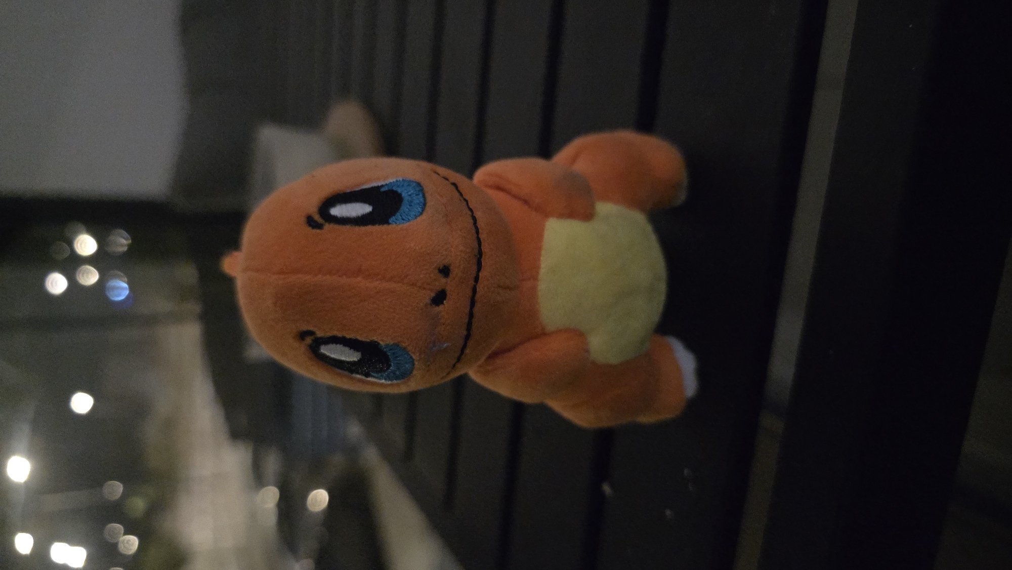 A close-up, slightly low-angle, eye-level shot features an orange plush toy of the Pokémon Charmander sitting on a dark slatted surface outdoors at night. The toy is centrally positioned, facing the viewer. It has large blue eyes with black pupils, a black embroidered mouth and nostrils, and a yellow belly. Its small orange arms are held close to its body, and its feet are visible at the bottom with white accents. The background is softly blurred, showing out-of-focus lights that suggest an urban or suburban setting at night. The slatted surface appears to be part of a table or bench. The overall lighting is dim, emphasizing the toy against the darker environment. This image was captured in Gan Yavne, Israel.