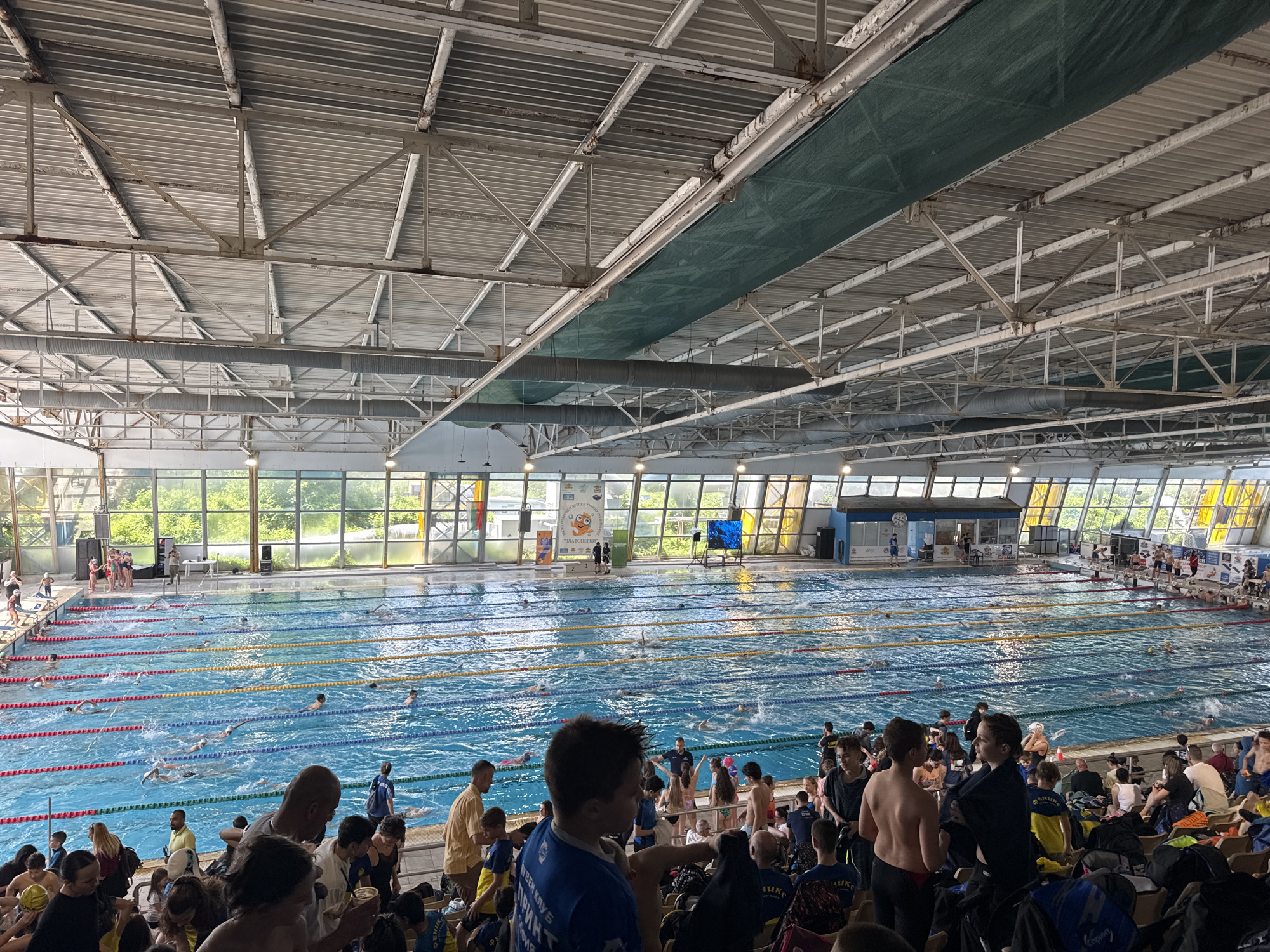 The image shows an indoor swimming pool complex filled with people. There are swimmers in the pool, which is divided into lanes marked by ropes. A crowd of spectators, including young people in athletic wear, are seated in the foreground. The background features large windows that provide natural light. The structure has an industrial-style roof with metal beams. It is the Vazrazhdane Swimming Complex in Sofia, Bulgaria.