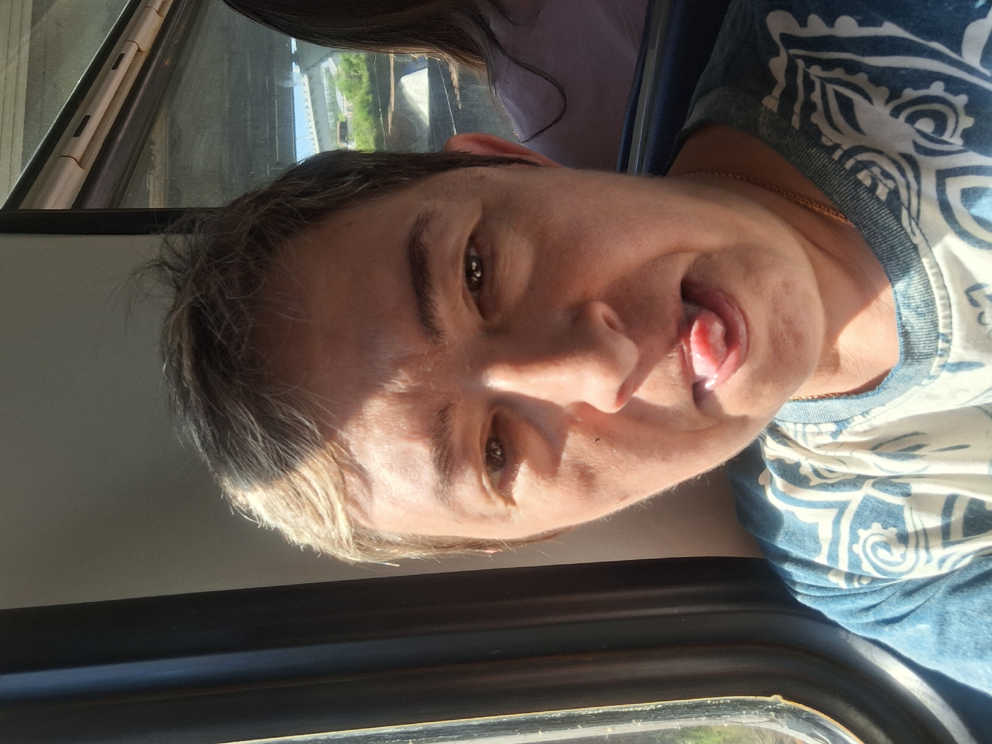 The image is a close-up, selfie-style photograph taken indoors, possibly on a train or bus, given the window on the left with a curtain and the reflection of scenery in the window on the right. A person with short, light brown hair and fair skin is the primary subject, positioned in the foreground and facing the camera directly. The person's tongue is sticking out, and their mouth is slightly open. They are wearing a blue t-shirt with a white, patterned design. Sunlight is hitting the left side of the person's face, casting shadows. In the background, through the right-side window, a bridge and greenery are visible, suggesting an outdoor environment. Another person, with dark hair and wearing a light purple shirt, is partially visible in the background on the right.