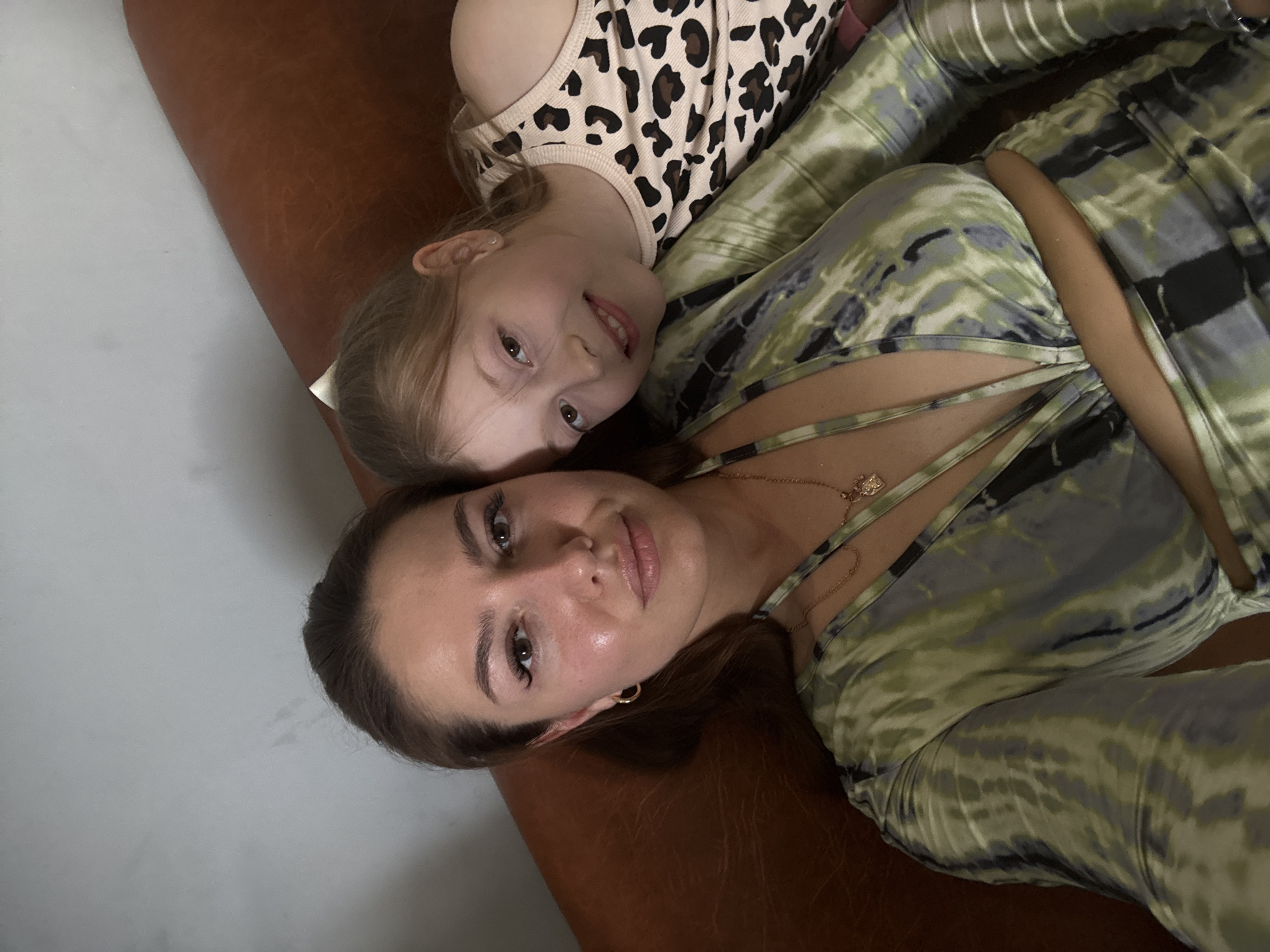 Two people, a woman and a young girl, are lying down, facing the camera. The woman is on the bottom, and the girl is lying on top of her, with her head near the woman's. Both are looking at the camera and smiling. The woman is wearing a green and gray tie-dye outfit, and the girl is wearing a white tank top with a leopard print. They appear to be on a couch or a similar surface, which is brown and textured. The background is a light gray wall. The lighting suggests it could be daytime, but no specific time can be determined. The overall mood is warm and affectionate, indicating a close relationship between the two individuals.