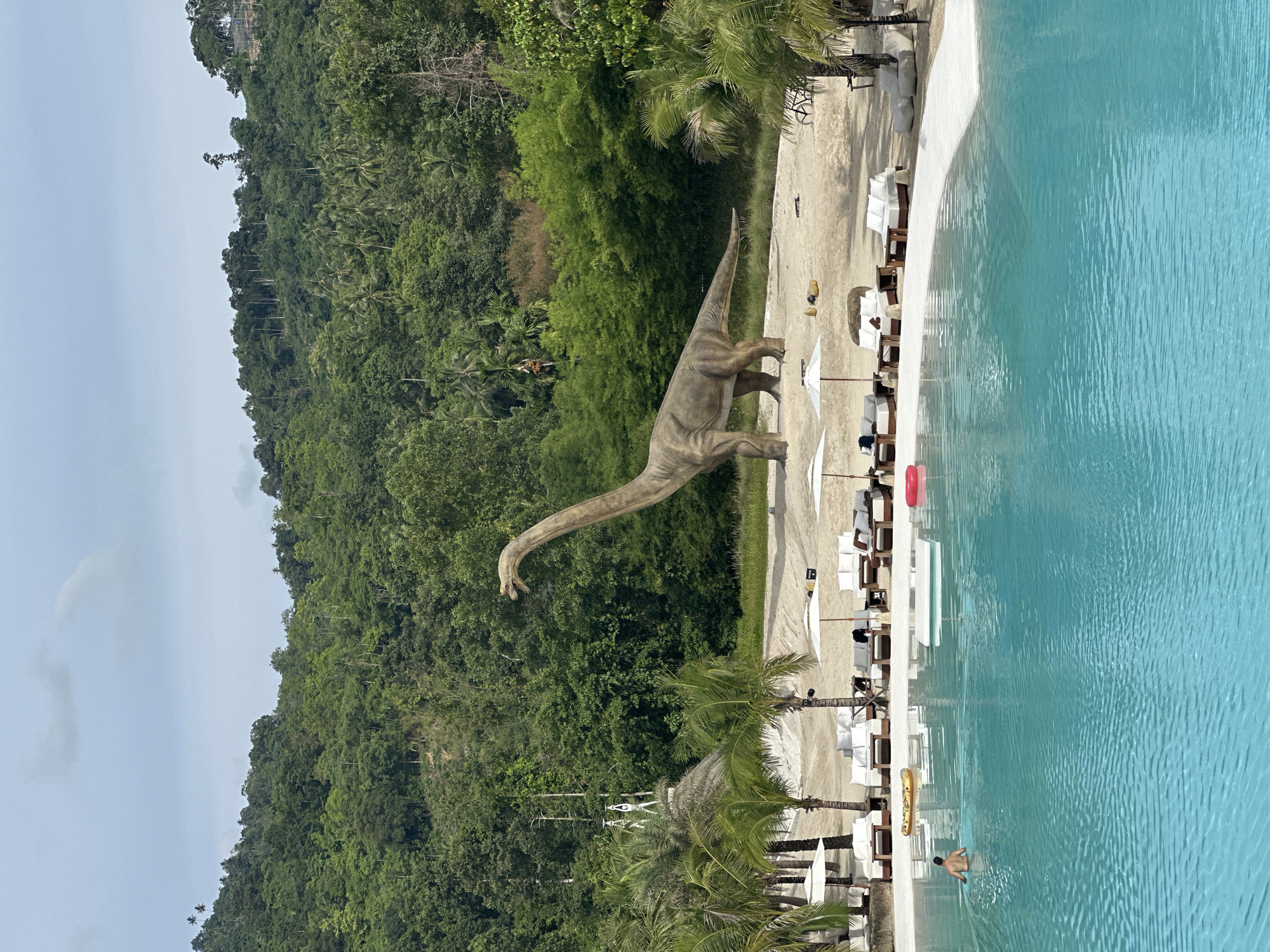 This image depicts a resort in Baan Lipa Noi, Thailand. In the foreground, a person swims in a bright turquoise pool. In the mid-ground, a sandy beach is lined with white lounge chairs and umbrellas. A large dinosaur statue stands on the beach, facing left. Palm trees and lush green hills covered in tropical vegetation form the background. The sky is partly cloudy and overcast.