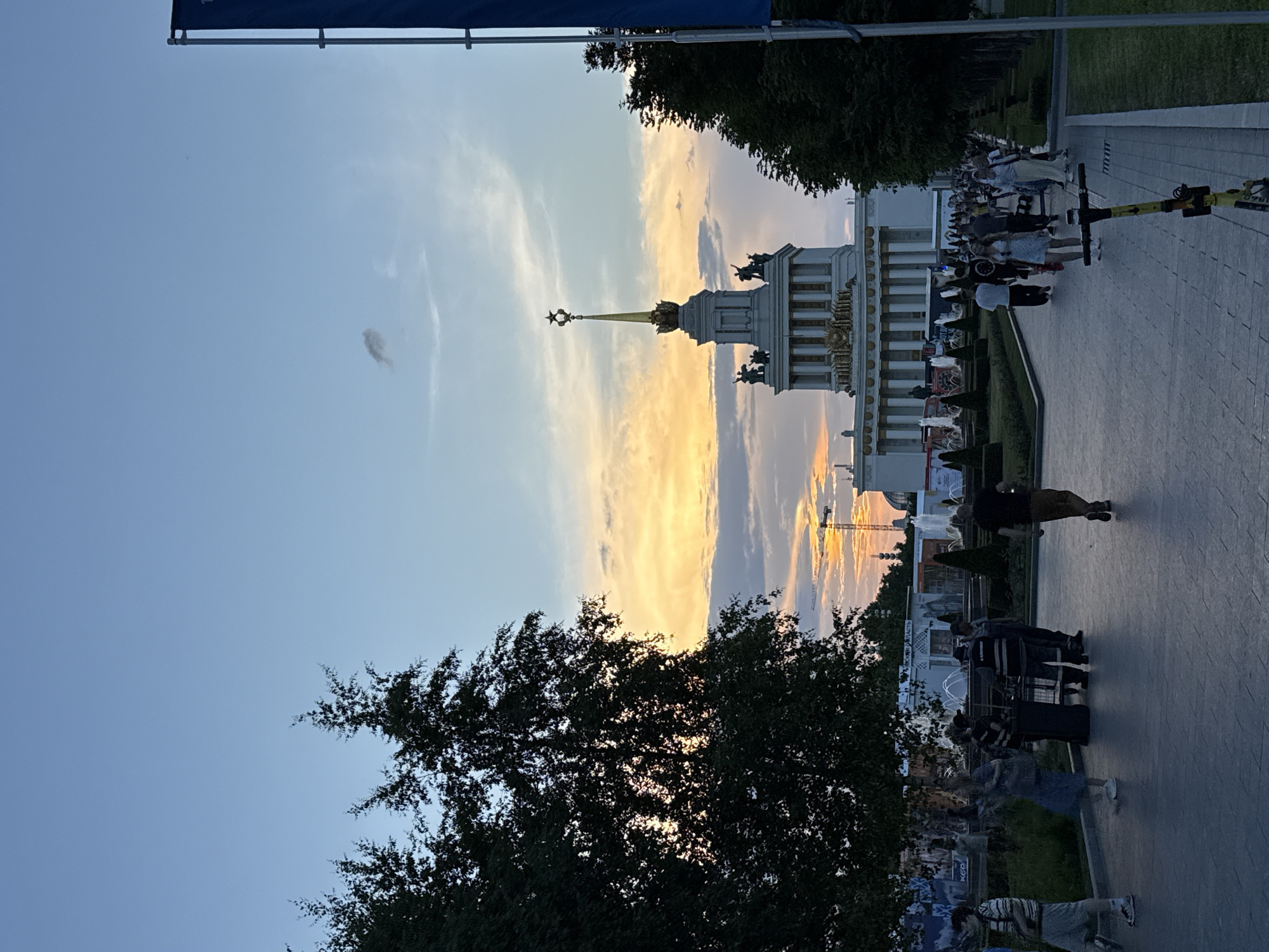 The image depicts the VDNKh (Exhibition of Achievements of National Economy) park in Moscow, Russia, during a sunset. A grand Soviet-era pavilion with a prominent star-topped spire stands in the background, illuminated by the fading sunlight. The sky above transitions from a clear blue at the top to hues of orange and yellow near the horizon, with scattered clouds.
In the foreground, a paved walkway is lined with trees on the right side, and green lawns with formal hedges and fountains on the left. Several people are walking along the pathway, some individually, others in small groups. Their clothing suggests warm weather. There are also some service workers, possibly park staff, visible near a fountain area. A yellow electric scooter is parked on the right side of the walkway. In the distance, a construction crane can be seen, indicating ongoing development or maintenance within the park. The overall atmosphere is one of a public space being enjoyed by visitors during a pleasant evening.