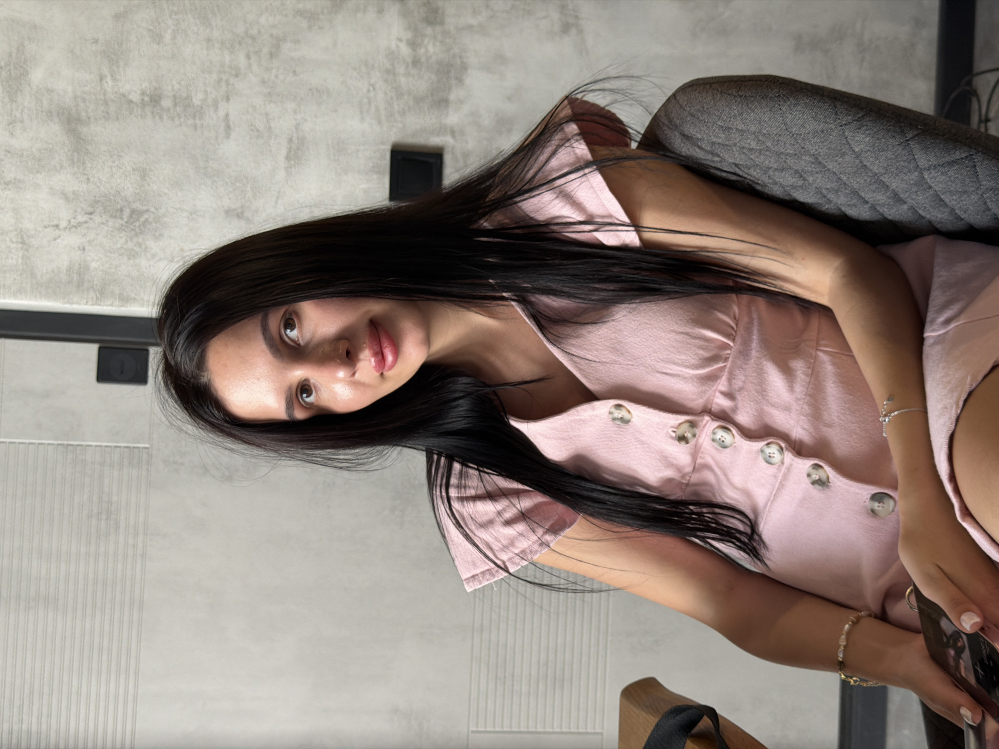 A young woman with long, dark hair and light skin sits in a chair in an indoor setting, possibly a cafe or restaurant. She is wearing a light pink dress with buttons down the front and has two bracelets on her left wrist. The background is a textured wall. The lighting suggests an indoor environment, likely during the day.