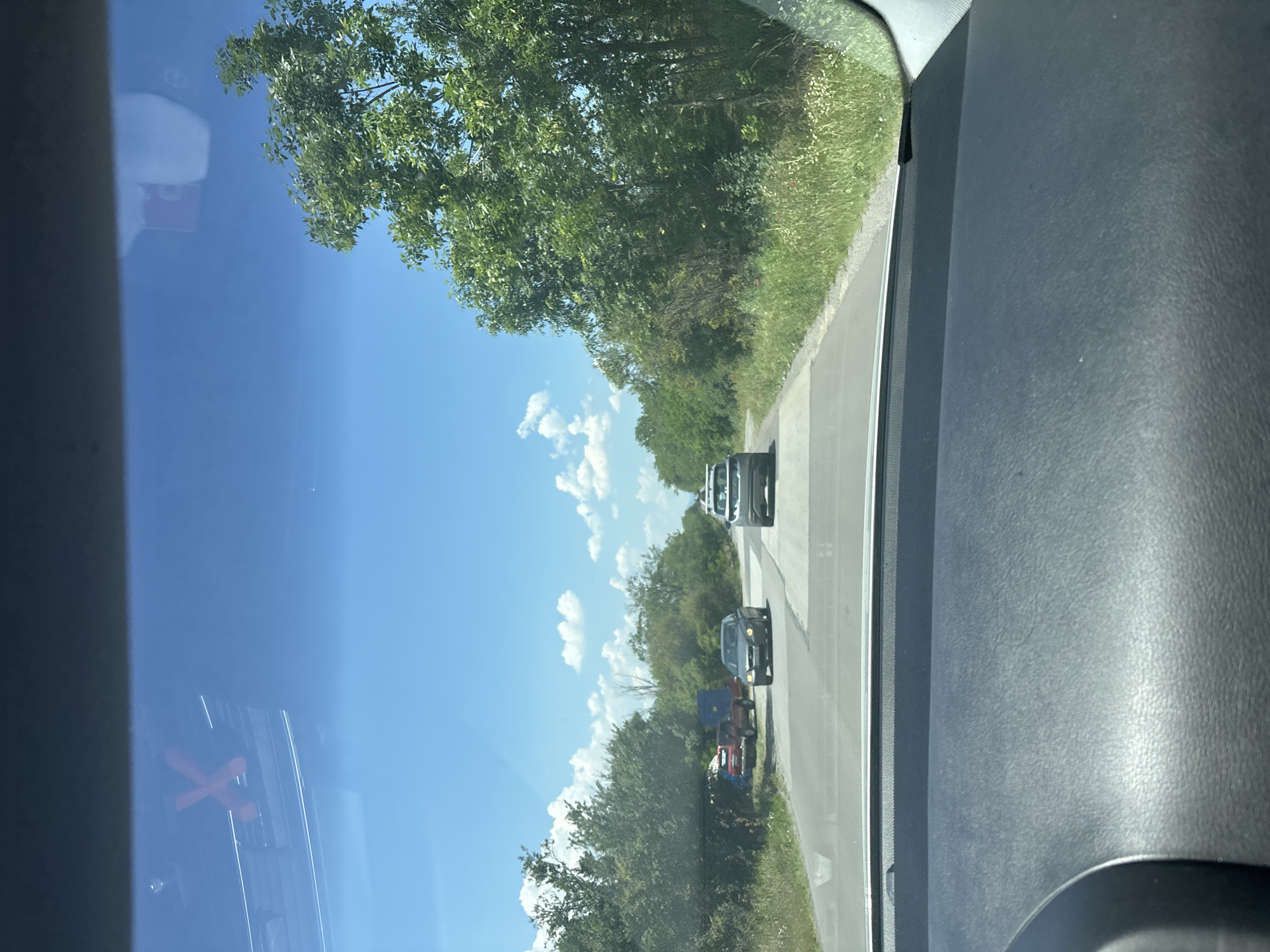 The image, taken from inside a vehicle looking forward, shows a two-lane road with cars in motion. The sky is blue with scattered white clouds. Lush green trees and vegetation line both sides of the road. On the left side, there is a red pickup truck towing a blue trailer parked near a cluster of trees. A dark-colored sedan is driving on the road, and a silver SUV is ahead of it. The dashboard of the vehicle from which the photo is taken is visible in the foreground. The scene appears to be a rural or suburban area, possibly on the outskirts of Lozen, Bulgaria, given the natural setting and the presence of the parked truck and trailer which could suggest a residence or temporary encampment.