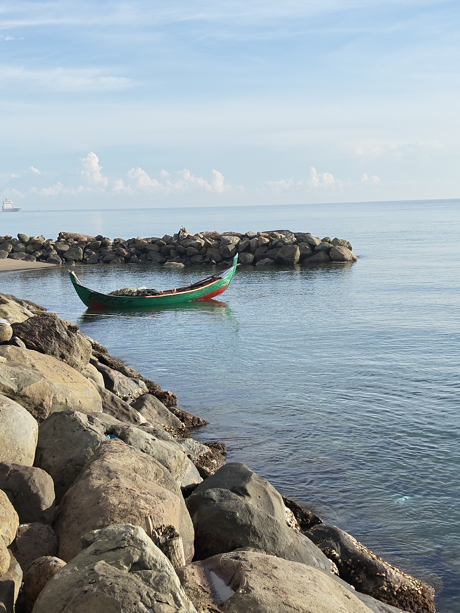 A medium-angle shot depicts a seascape in Lhokseumawe, Indonesia. A green and red boat floats in the water, secured to rocks. A breakwater of rocks extends into the ocean, with two figures visible on the rocks. Large boulders are in the foreground. The water is calm and clear. The sky is pale blue with scattered white clouds. A distant ship is visible on the horizon.
