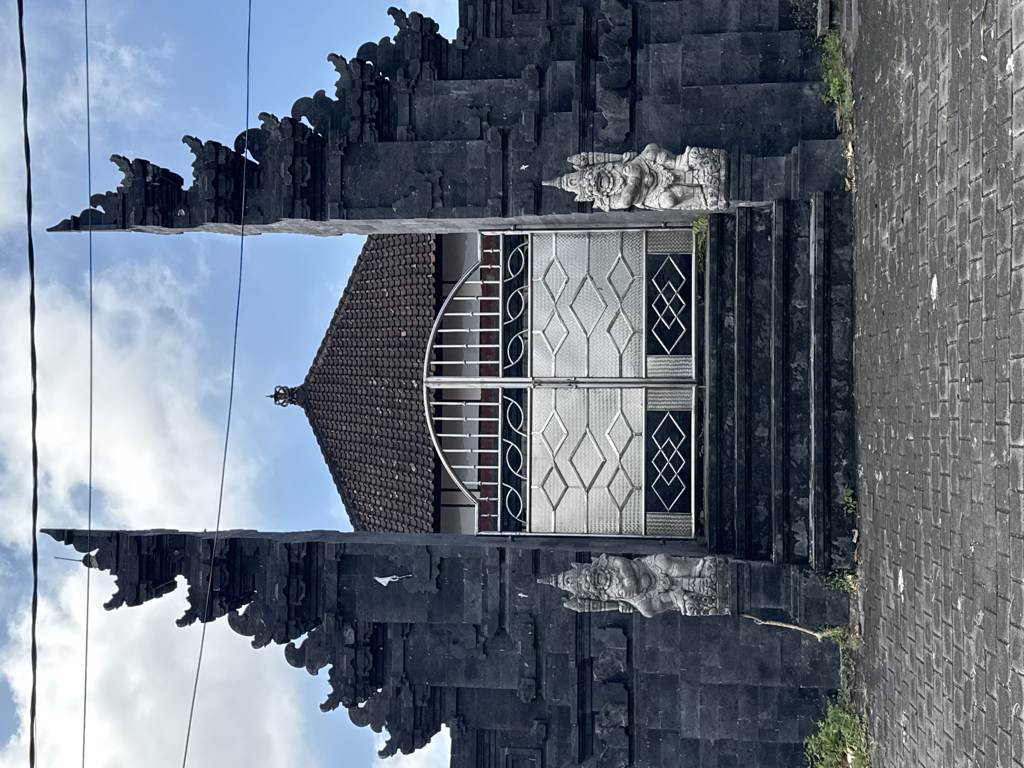 The image captures the entrance to a building in Kuta Selatan, Indonesia. The entrance is framed by a black stone structure with intricate carvings, characteristic of Balinese architecture. Two stone statues stand guard on either side of the entrance. A set of stairs leads up to a metal gate with a geometric design. Behind the gate, the roof of a building is visible. The sky is visible through the opening. The ground is paved with gray brick. Power lines are visible in the top left corner.