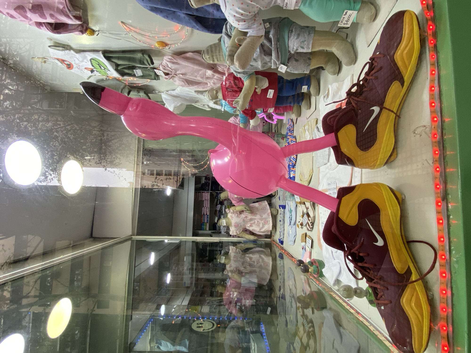 The image displays a retail store window in Buenos Aires, Argentina, featuring a children's clothing display. Central to the foreground is a large, upright pink flamingo figure, likely plastic or inflatable, with its two leg-like protrusions inserted into a pair of maroon and yellow Nike athletic shoes. These shoes feature white Nike swooshes and are positioned side-by-side on a green-edged display surface, which is illuminated along its front edge by a strip of red LED lights.
Behind the flamingo, several child mannequins, some with featureless fabric-covered heads and others headless, showcase various items of baby and children's apparel. Noteworthy examples include two mannequins wearing pink floral dresses with ruffled sleeves, and others displaying outfits such as a red hooded top with blue pants, a grey patterned top and shorts, and a white top with a green alligator graphic. Additional folded or laid-out clothing items, including baby onesies and blankets, are scattered across the display surface. Decorative elements include small figurines, glowing crescent moon and star string lights, and small circular yellow lights integrated into the display.
Reflections on the glass storefront reveal elements of an urban environment, including tree branches and multi-story buildings, indicating the display faces a street. Interior lighting fixtures, appearing as bright white circles, are visible both directly and as reflections. The store's interior, visible behind the display, shows shelves stocked with neatly folded textiles and clothing items. A yellow smiley face is reflected on the left side of the window. Price tags, one showing "$10000," are visible on some displayed items.