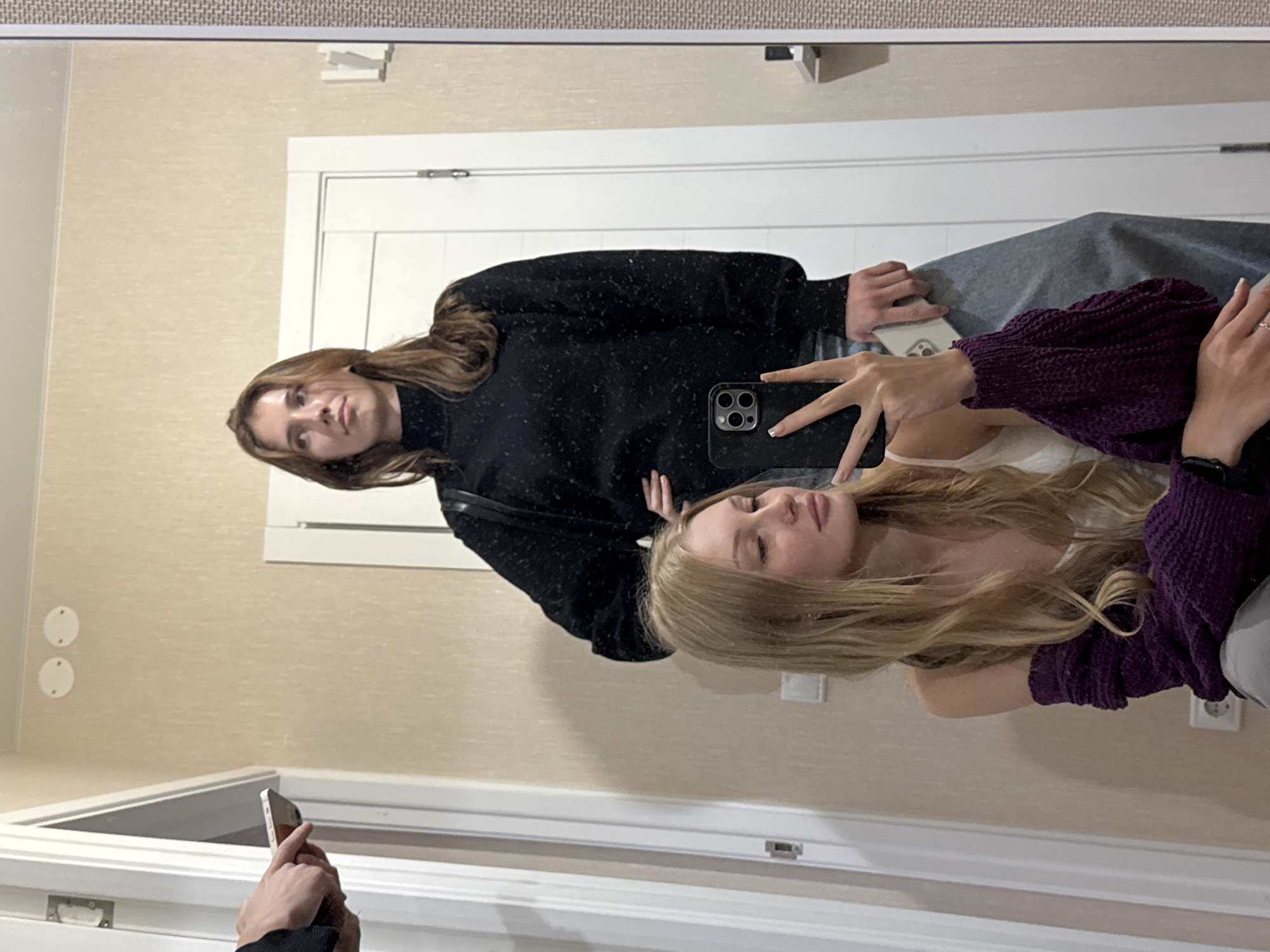 Two women are reflected in a mirror. The woman in the foreground has long blonde hair and is wearing a white top and a purple knitted sleeve. She is holding a smartphone and taking a selfie. The woman behind her has long brown hair and is wearing a black turtleneck sweater and gray pants. She is looking to the side. In the background, there is a white door and a beige wall. A hand holding a smartphone is visible on the left side of the image. The environment appears to be an indoor hallway, possibly in an apartment.