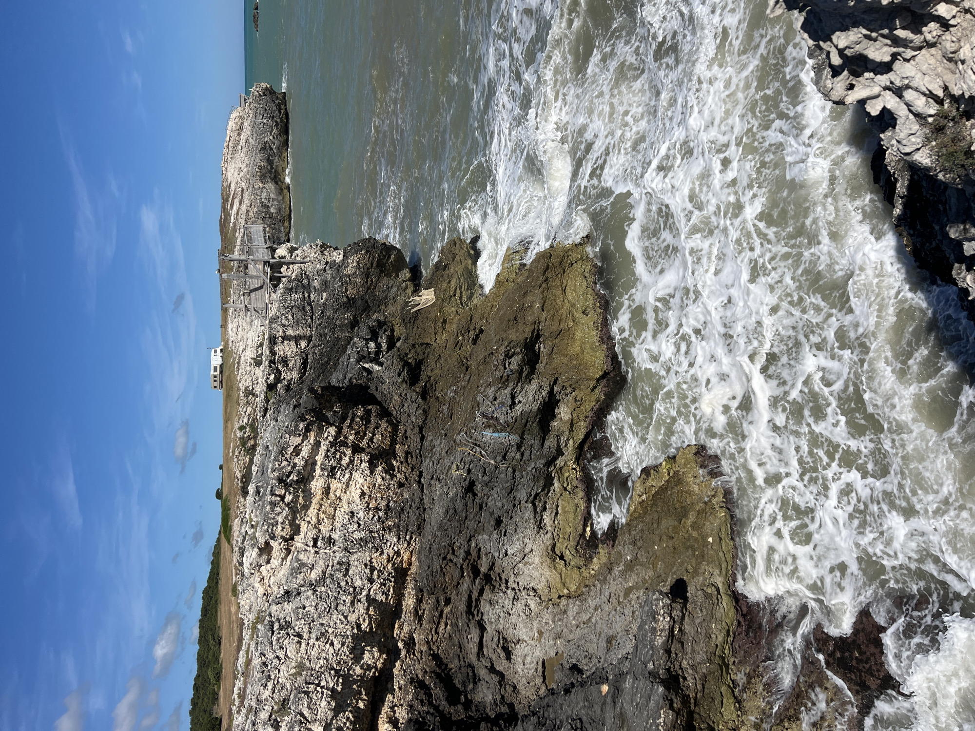 A rugged cliffside meets the choppy, greenish-gray waters of the Adriatic Sea under a bright blue, partly cloudy sky. The cliffs are layered with rock formations in shades of gray, white, and brown, with patches of green moss and vegetation clinging to the surfaces. Wooden walkways with railings are constructed along the cliff edge, providing access to viewing areas. In the distance, a white recreational vehicle is parked on a grassy plateau overlooking the sea. The water is agitated, with white foam and waves crashing against the rocks at the base of the cliffs. The scene is likely located in the coastal region of Peschici, Italy, known for its dramatic limestone cliffs and clear waters.
