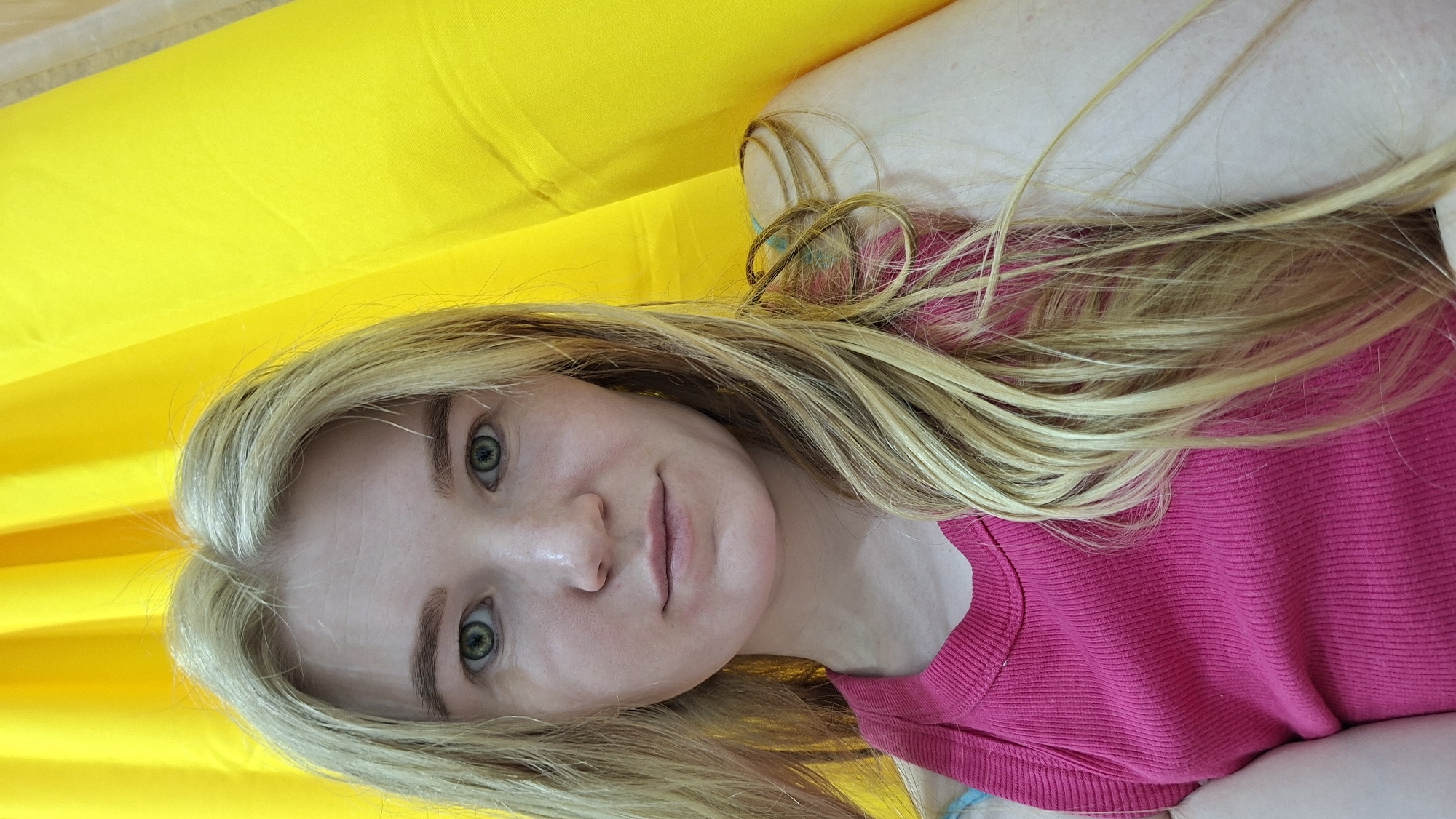 This portrait photograph features a fair-skinned woman with long, wavy blonde hair and striking light green or hazel eyes. Her lips are a natural light pink, and her expression is calm and direct as she looks into the camera. She appears to be in her twenties or thirties.
She is wearing a vibrant fuchsia or hot pink, ribbed knit top with a crew neck. The image is oriented sideways, with her head positioned on the left and her body extending to the right. She appears to be lying down, perhaps relaxing.
The woman is set against a bright, solid yellow background, which seems to be a soft fabric surface, possibly a large pillow or blanket. A lighter, almost skin-toned or white fabric surface, is also visible behind her head on the right side of the frame. The lighting is bright and even, suggesting a daytime setting indoors, either with natural light or good artificial illumination. There is no specific activity depicted other than posing for the photograph. No text is visible in the image. While the image location is contextually given as Chaikovsky, Russia, there are no visual cues within the frame to confirm this.