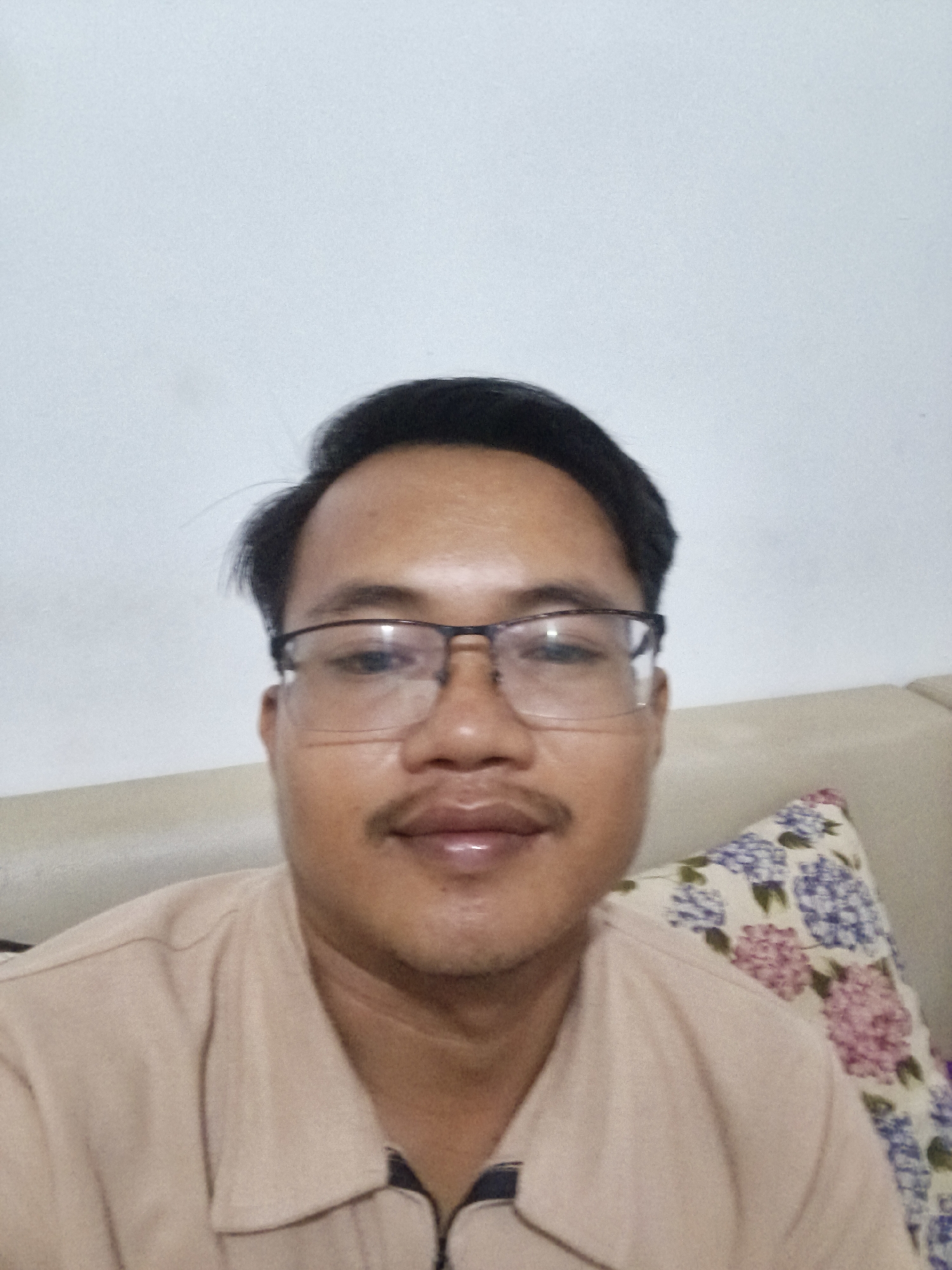 A medium shot selfie taken from a slightly low angle shows a man with dark hair, a mustache, and glasses. He is wearing a light brown collared shirt. He is positioned in front of a plain white wall, with his right shoulder resting against a beige sofa with a floral patterned pillow. The background suggests an indoor setting, possibly a residence in Pangkalpinang, Indonesia, though no specific landmarks are visible. The lighting is neutral, and the overall mood is casual and relaxed.