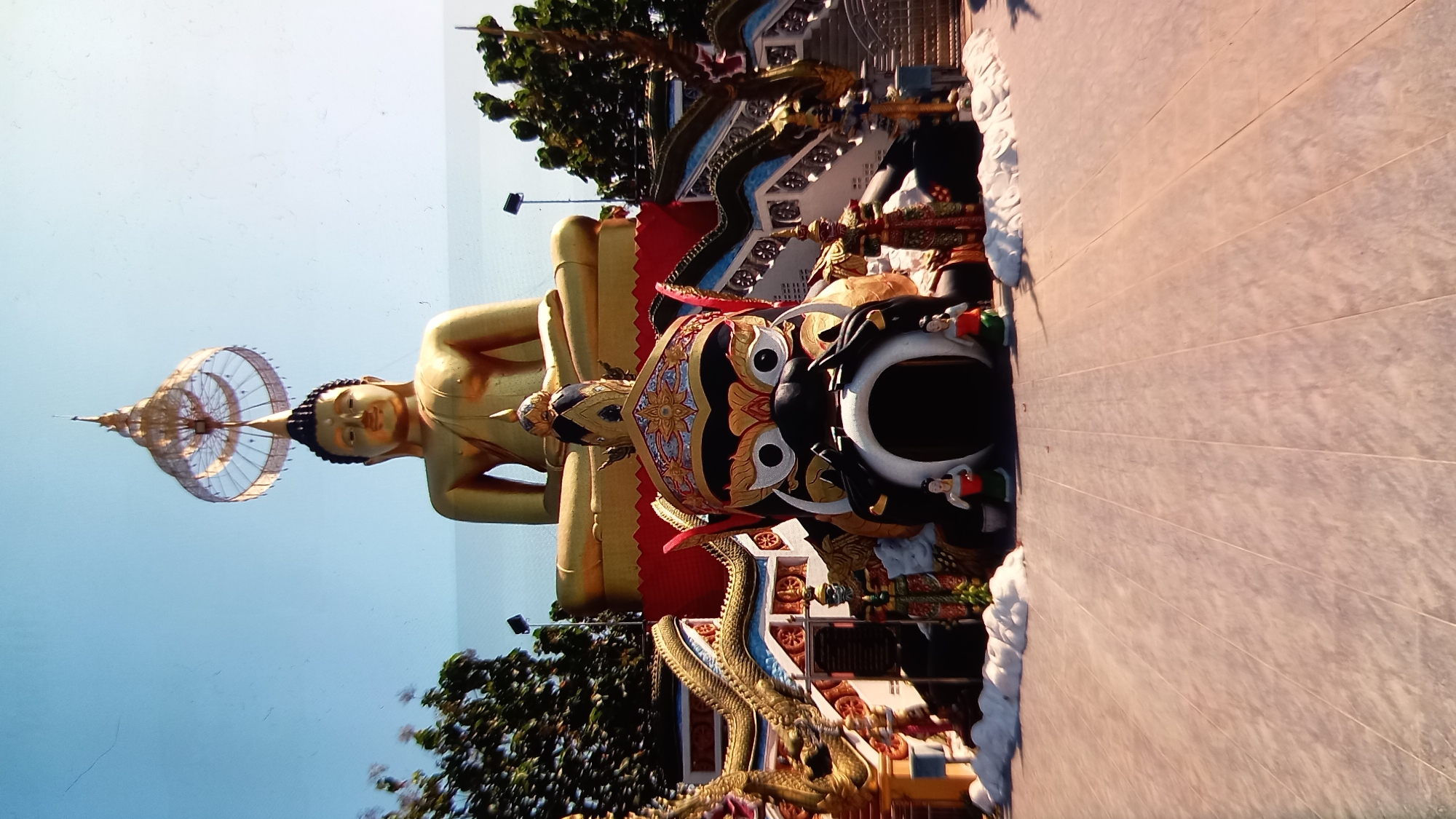 This media file depicts a large, golden statue of a seated Buddha with an elaborate, tiered headdress. The statue is positioned outdoors, with a clear blue sky as the backdrop. In front of the Buddha, there is a large, dark-colored statue with a fearsome face, elaborate headdress, and large eyes, possibly a mythological guardian figure. A decorative, serpentine dragon sculpture is also visible near the base of the Buddha. The ground in front of the statues is paved with light-colored tiles. The scene appears to be in a temple or a similar religious or cultural site. The lighting suggests it is daytime, possibly late afternoon due to the warm tones on the tiled ground. The overall atmosphere is serene and majestic, typical of Buddhist iconography. No visible text is present in the image. The location context provided is Ko Phangan, Thailand, which aligns with the visual cues of tropical foliage and possibly Southeast Asian religious art.