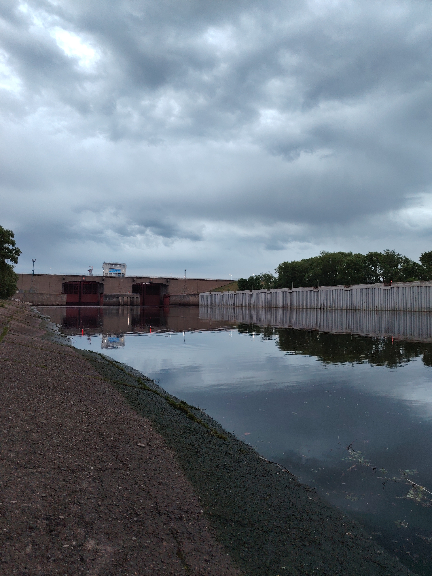 The image shows a view of the Kama River and the dam in the city of Chaikovsky, Russia, under a cloudy sky. The river is in the foreground, with a concrete embankment on the left. In the background is the dam structure, with red gates and a building with a sign in Russian. Trees line the right side of the river behind a concrete wall. The water reflects the sky and structures.