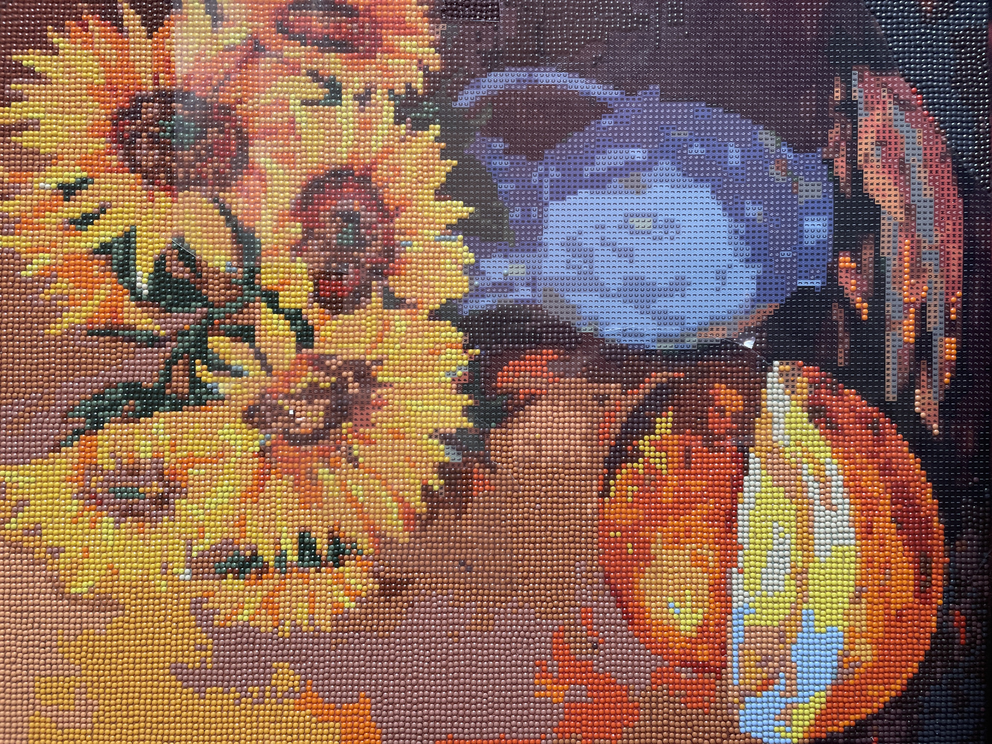 This is a close-up, high-angle shot of a diamond painting depicting a still life of sunflowers in a vase, a bowl of fruit, and a saucer. The painting is rendered in small, faceted beads of various colors, giving it a textured, pixelated appearance. The sunflowers are dominant in the upper portion of the image, with vibrant yellow petals and dark brown centers. To the left, a bright orange bowl overflows with a variety of fruits, primarily depicted in shades of yellow, orange, and white, with a few accents of light blue. To the right of the fruit bowl sits a blue pitcher, its surface adorned with a swirling pattern in lighter shades of blue and white. The background is a deep, mottled brown. The diamond painting is likely a commercial art piece and not indicative of any specific location within Shpykolosy, Ukraine, or any other city.