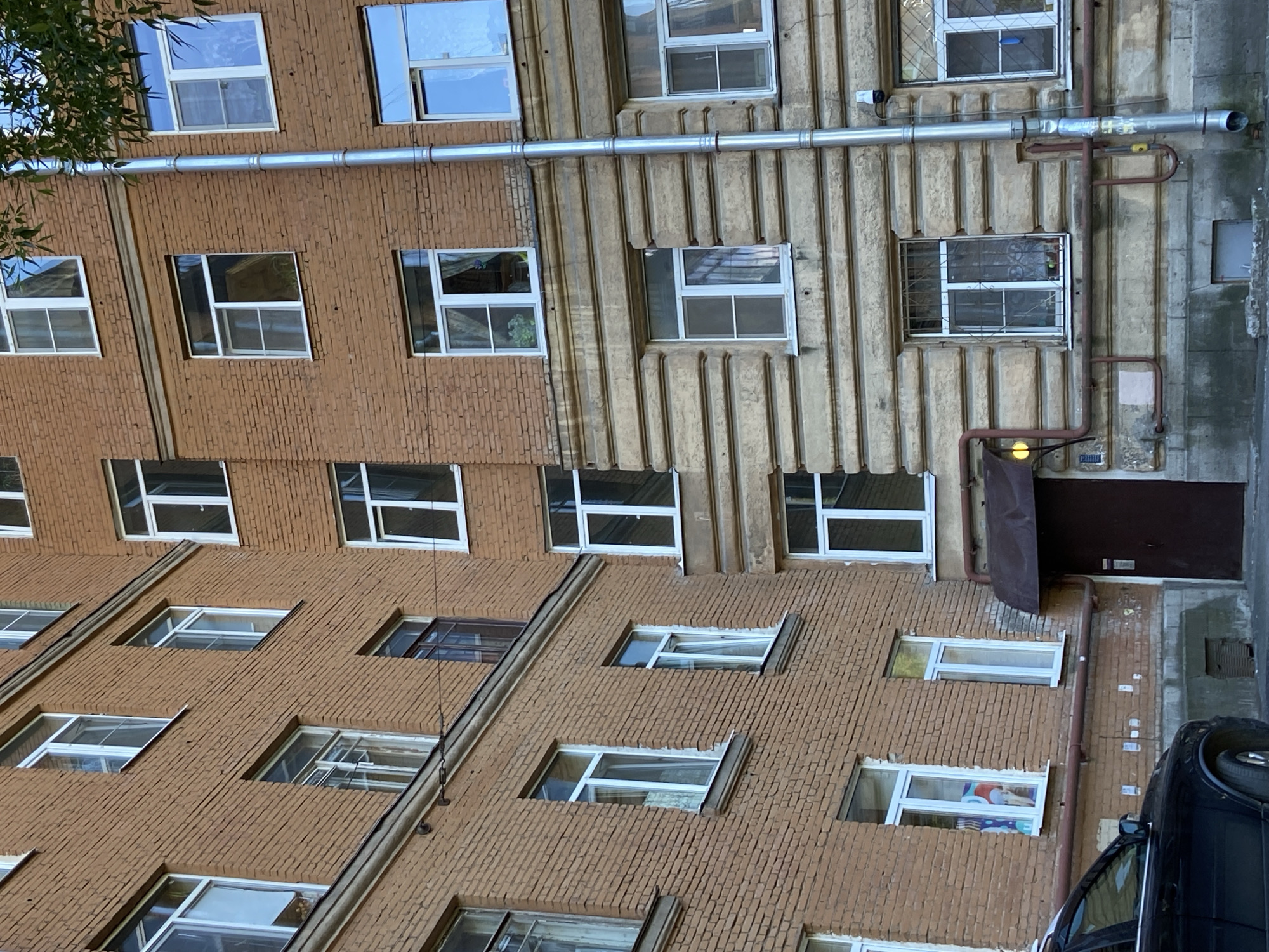 The image depicts the exterior of an old brick apartment building in Saint Petersburg, Russia. The building is constructed with reddish-brown bricks and features multiple windows, many with white frames. The lower section of the building has a facade with horizontal, stone-like detailing. A silver downspout runs vertically along the right side of the building. A dark brown door is visible on the ground floor, partially covered by a dark awning. A black car is parked in front of the building on the left side. The sky is visible at the top, with some tree branches with green leaves overhanging the building. The overall impression is of a typical residential building in an older urban area.