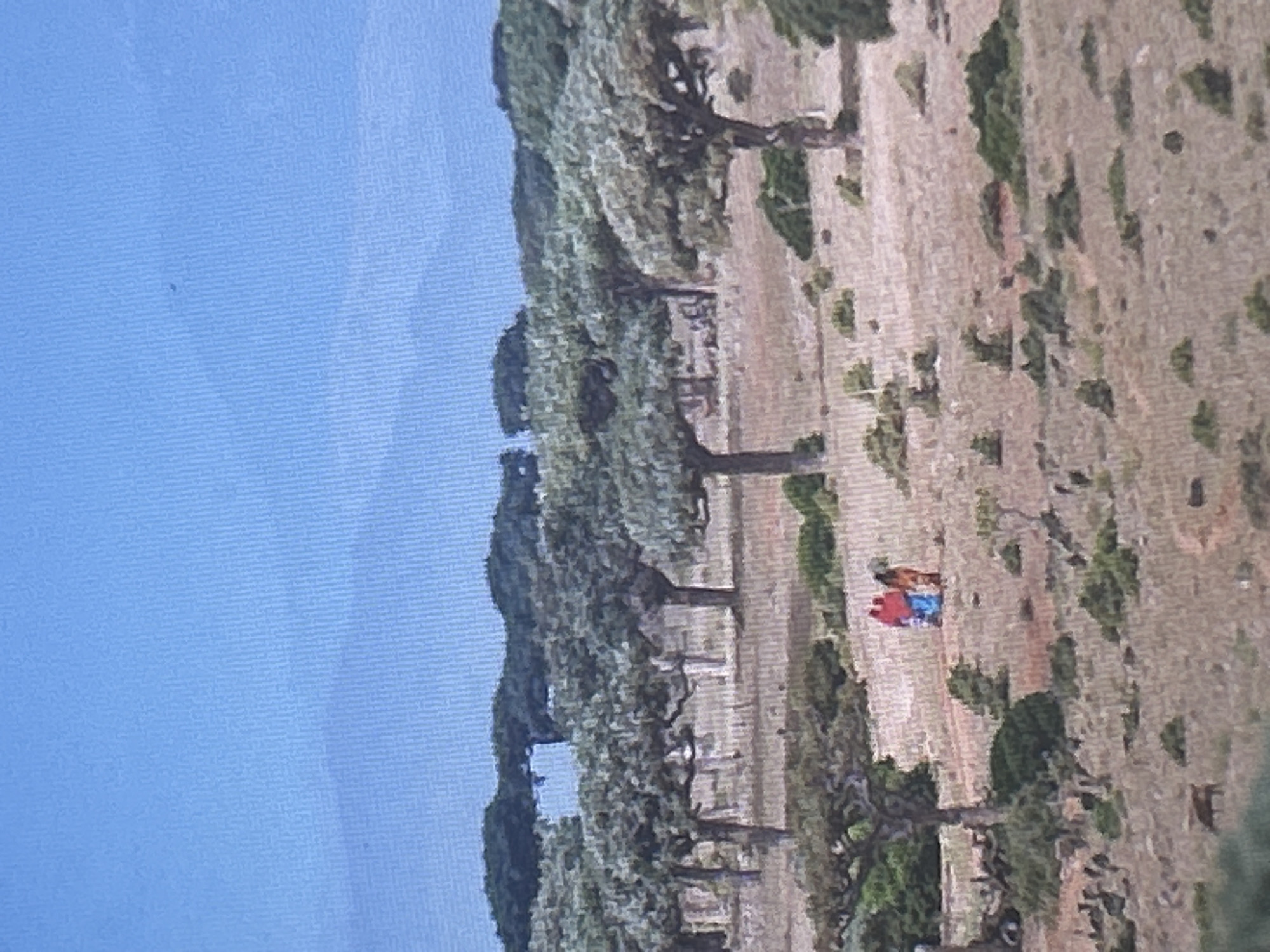 The image depicts a dry, sparse landscape with scattered, gnarled trees, possibly a desert or arid region. In the middle ground, two figures are walking on a dirt path. One is wearing a bright red garment, and the other is in an orange and blue garment. The ground is covered in dry grass and scattered shrubs. In the background, there is a range of hills or mountains under a clear blue sky. There is no indication of the city of Sofia, Bulgaria, in this image.