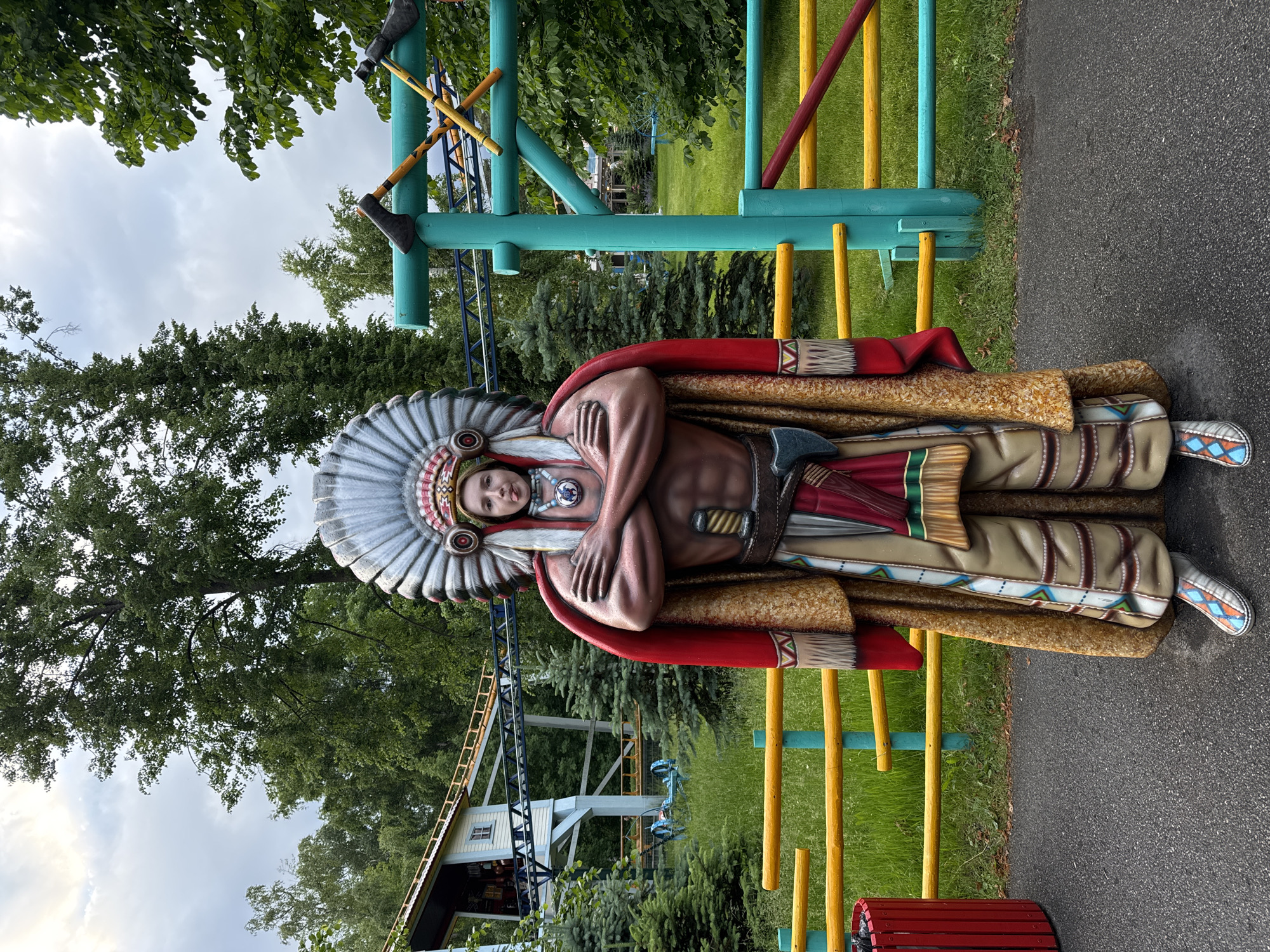A full-body statue of a person dressed as a Native American is the central focus of the image. The statue is positioned in what appears to be an outdoor amusement park. The figure wears a large feathered headdress, a red and gold cloak, a loincloth, and intricately patterned leggings. It has its arms crossed over its chest and appears to be holding a tomahawk.
In the background, a roller coaster track is visible, suggesting the location is an amusement park. There are also trees and greenery, indicating an outdoor setting. A colorful fence, made of yellow, blue, and red horizontal bars supported by turquoise posts, runs horizontally across the middle ground of the image. To the left of the statue, a red trash bin is present. The ground in front of the statue is paved with asphalt, while the area behind the fence is covered in grass. The sky is overcast with a cloudy sky. The statue's facial expression is a playful pout, with its tongue sticking out slightly.
The location is the "Divo Ostrov" amusement park in Saint Petersburg, Russia, known for its amusement rides and theming.