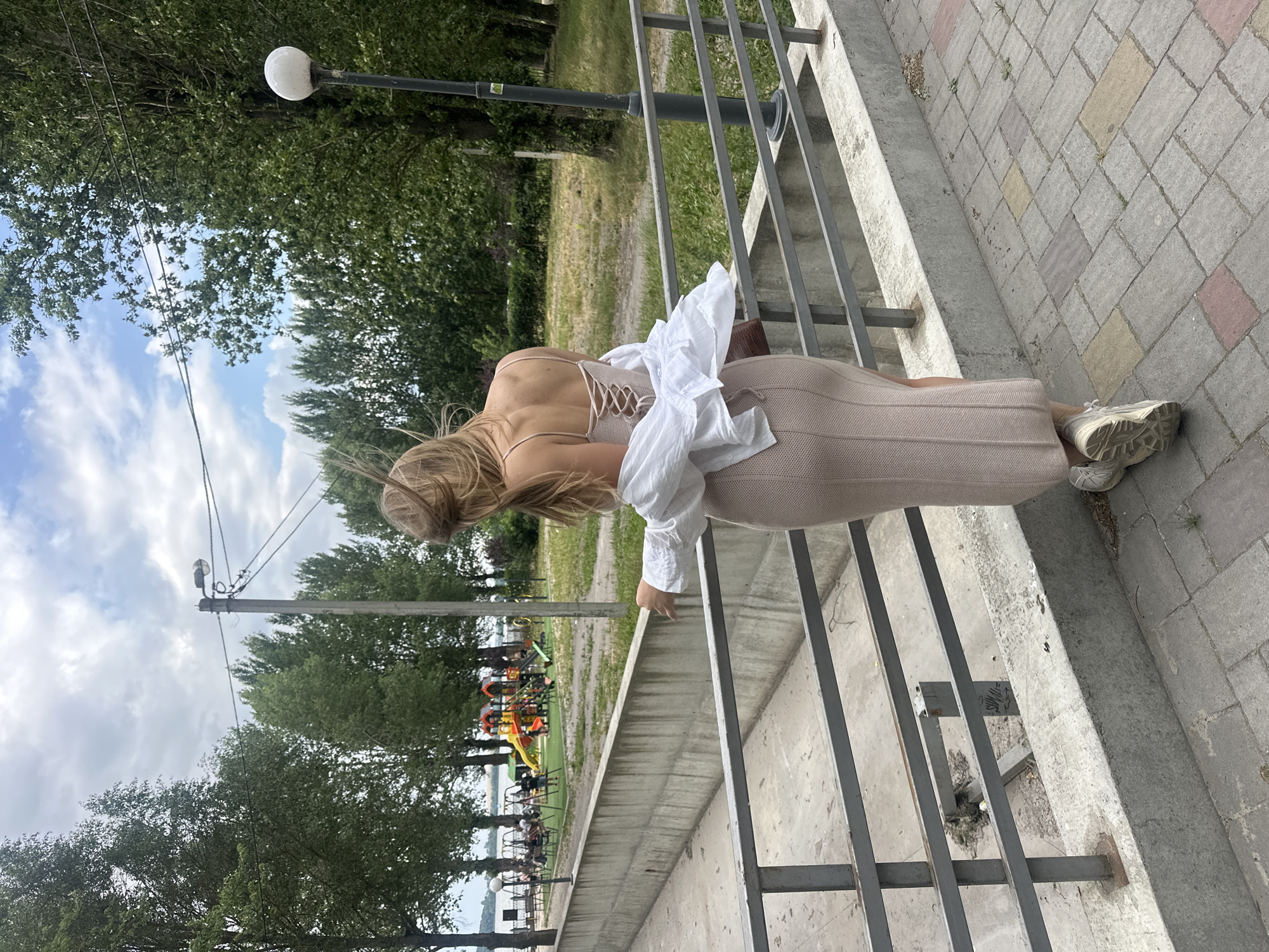 The image depicts a young woman from the back, standing on a paved walkway next to a metal railing. She is wearing a light pink knitted dress with a lace-up detail in the back and a white unbuttoned shirt over it. Her hair is blonde and appears windswept. She is looking out towards a park area with playground equipment, trees, and a body of water in the distance. The walkway is made of concrete and brick pavers, and there is a concrete retaining wall to her left. A lamppost is visible in the background. The location is likely a park or embankment along the Ternopil pond in Ternopil, Ukraine.