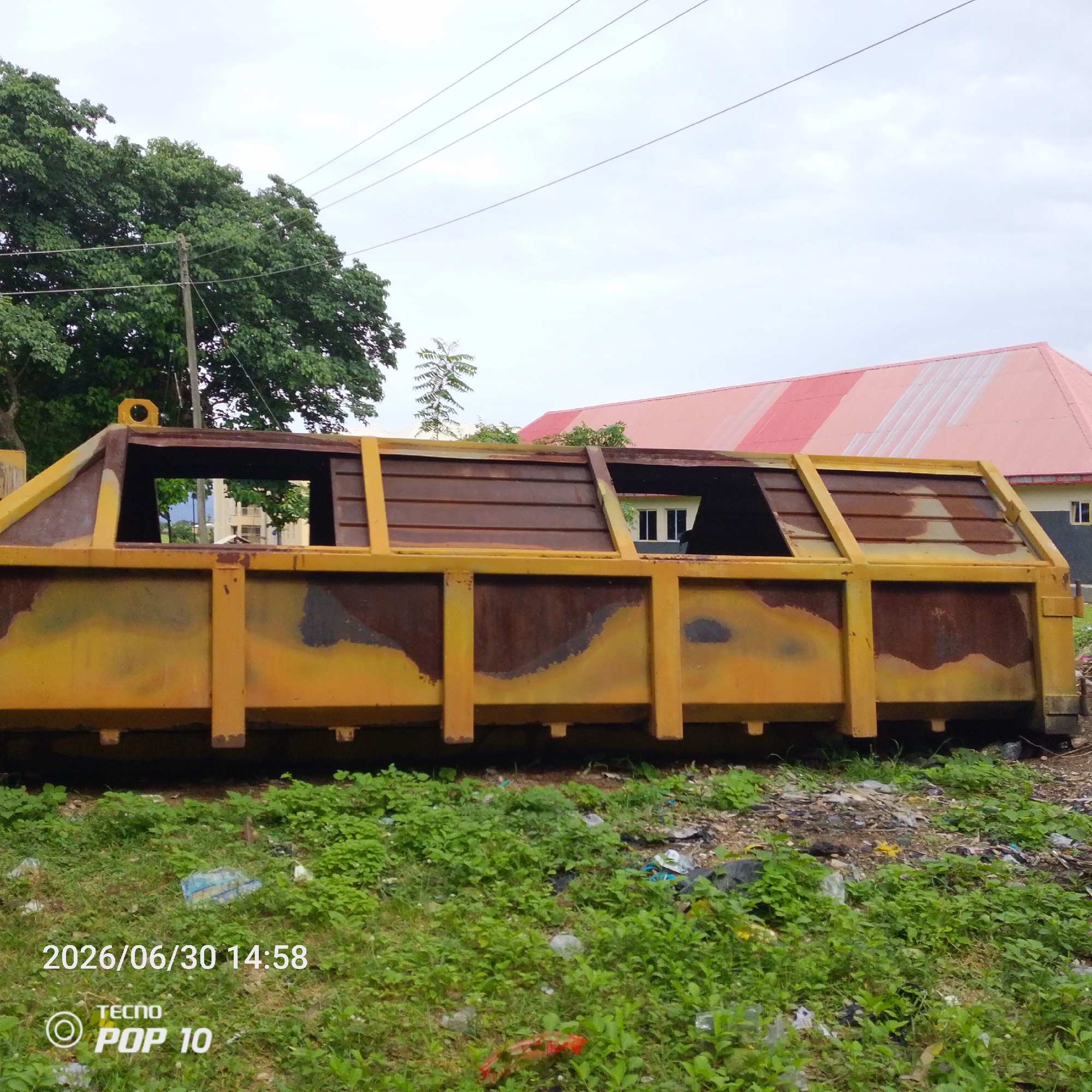 The media file shows a large, rusty, yellow skip or dumpster in Jalingo, Nigeria. The skip is empty and appears to be outdoors, situated on a patch of overgrown green grass and scattered debris. In the background, a building with a red, ridged roof is visible, along with some trees and utility poles. The sky is overcast, suggesting a cloudy or possibly rainy day. There is no activity or event taking place; the scene appears static.
Notable details include the weathered condition of the skip, with rust and faded paint evident, indicating it might be old or exposed to the elements. The surrounding area is somewhat untidy, with visible trash and weeds.
Visible text includes a timestamp in the lower left corner: "2026/06/30 14:58" and the text "TECNO POP 10" with a circular logo, likely indicating the device used to capture the image. The date suggests a future timestamp. The scene is set in Jalingo, Nigeria, offering a glimpse into an outdoor, possibly industrial or waste management area. The overall mood is neutral, depicting an ordinary outdoor scene.