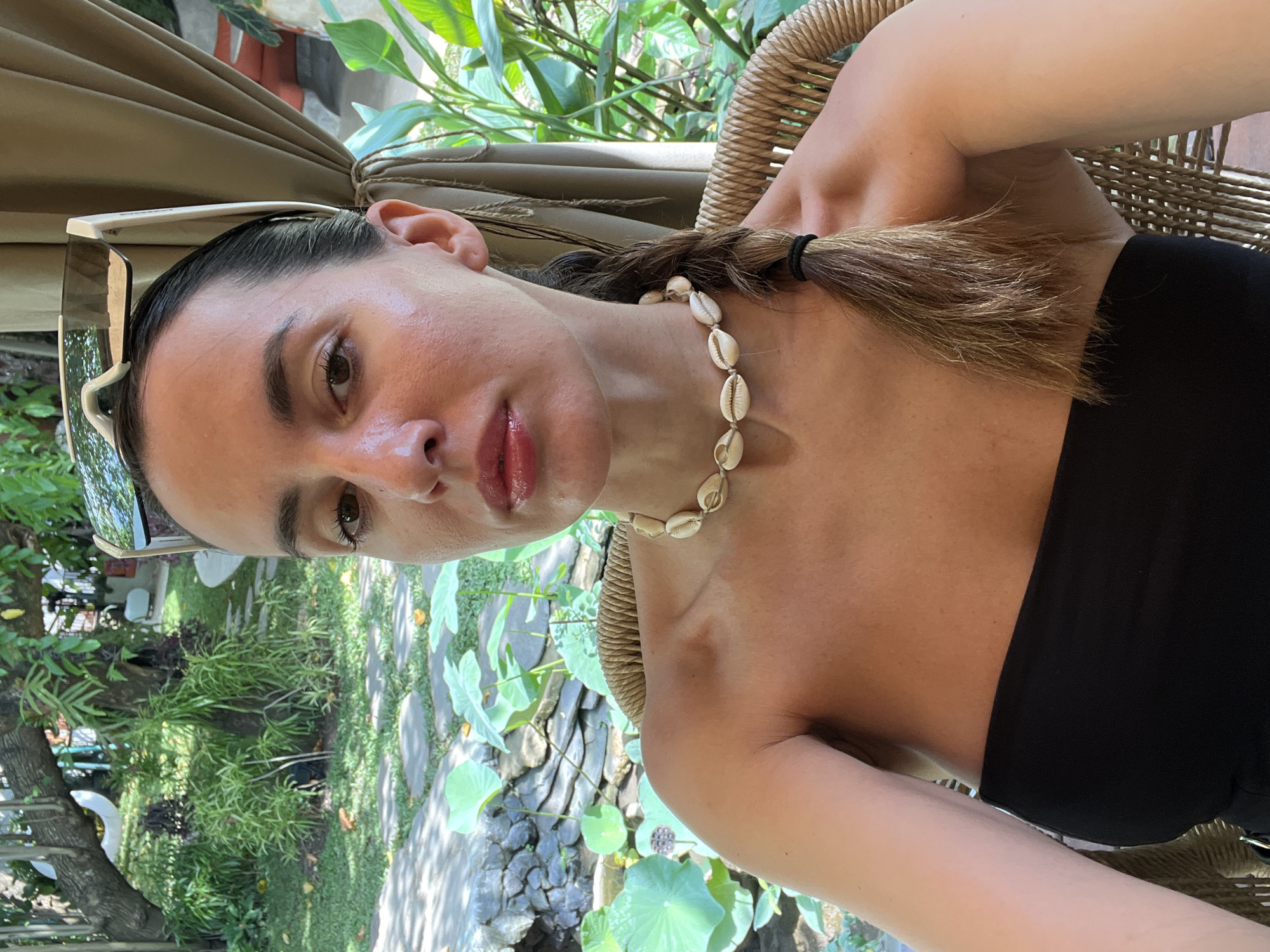 A medium, eye-level shot of a fair-skinned woman, probably in her late 20s or early 30s, wearing a black, strapless top and a necklace made of seashells. She is posed in a woven chair, possibly at an outdoor cafe or patio. Her dark hair is pulled back into a braid. A pair of sunglasses is hooked on her hair. She looks directly into the camera. The location appears to be North Kuta, Indonesia, as evidenced by the tropical vegetation and the style of the outdoor setting. In the background, there is a garden landscape, including a stone path, various green plants, and a pond featuring lotus pads. A beige-colored curtain or canopy can be seen overhead.