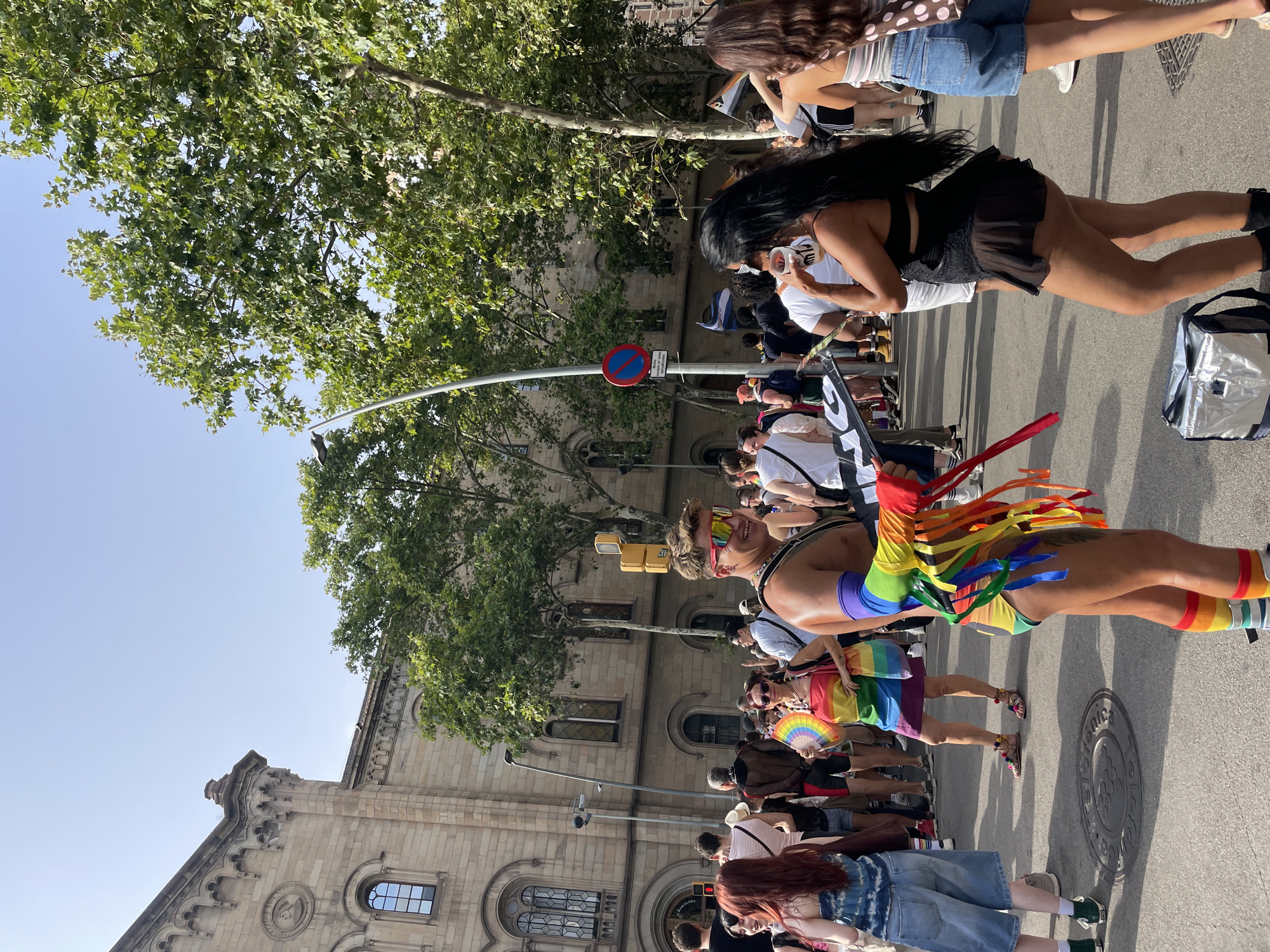 The image captures a vibrant street scene in Barcelona, Spain, likely during a Pride event given the prominent display of rainbow colors. A group of people are gathered on a paved street lined with historic buildings and lush trees. The architecture suggests a European city with ornate facades and arched windows.
In the foreground, a person with a muscular physique and wearing rainbow-striped armbands and socks, along with a colorful, fringed garment, is holding a sign. They are smiling and appear to be a participant in a parade or celebration. To their left, a woman with red hair is also dressed in a rainbow-colored outfit and holding a fan shaped like a rainbow.
Further back, a diverse crowd is visible, many of whom are casually dressed. The background shows a street with traffic lights and signs, indicating an urban environment. The presence of large trees suggests a well-established city with green spaces. The clear blue sky indicates a sunny day. The overall atmosphere appears festive and communal.