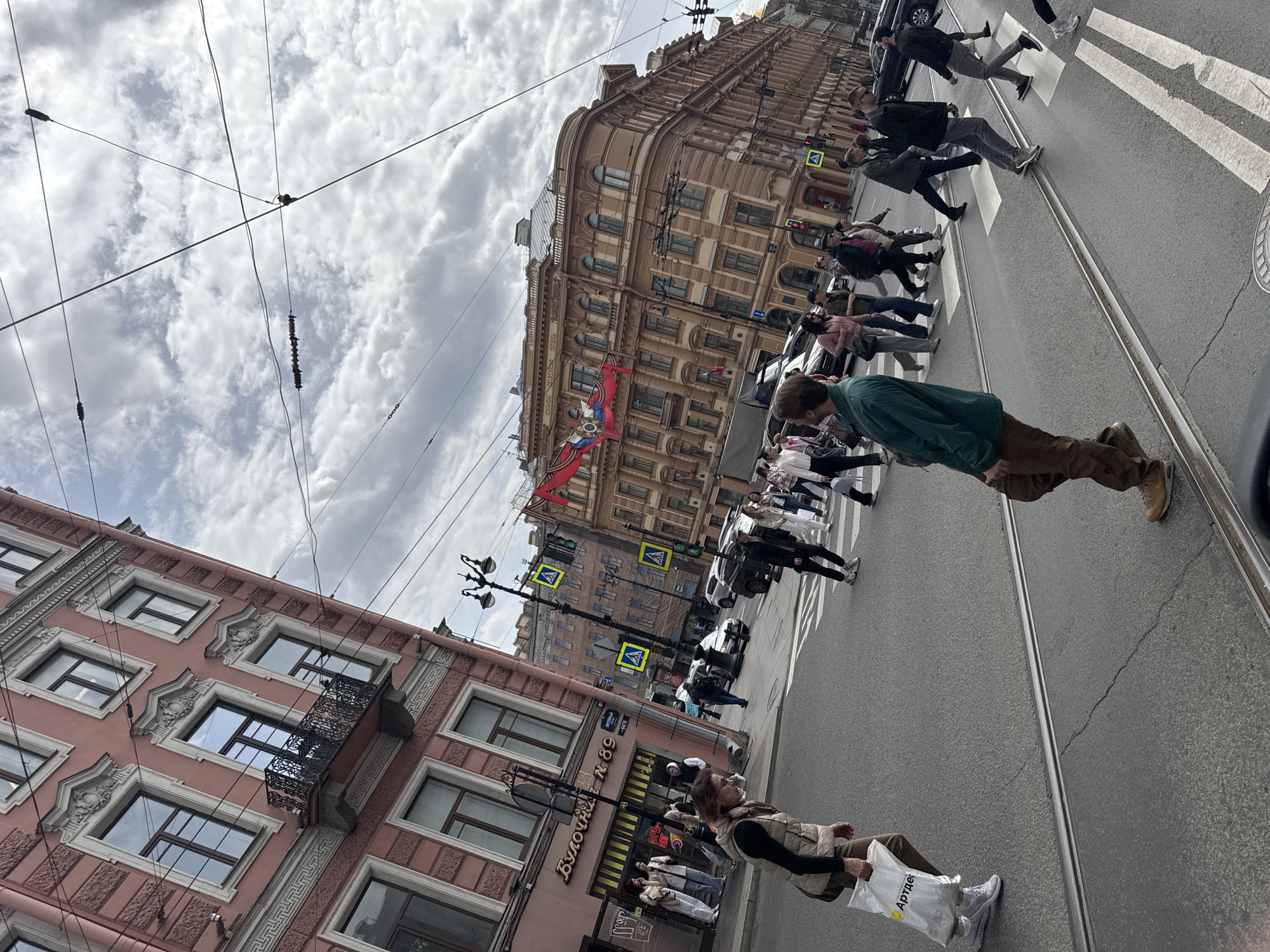 The image depicts a street scene in Saint Petersburg, Russia. People are crossing a crosswalk on a street with tram tracks. Buildings with detailed facades line the street, one with the sign "Булочная N 89" (Bakery N 89). A Russian flag hangs on one of the buildings. The sky is cloudy, and overhead wires are visible.