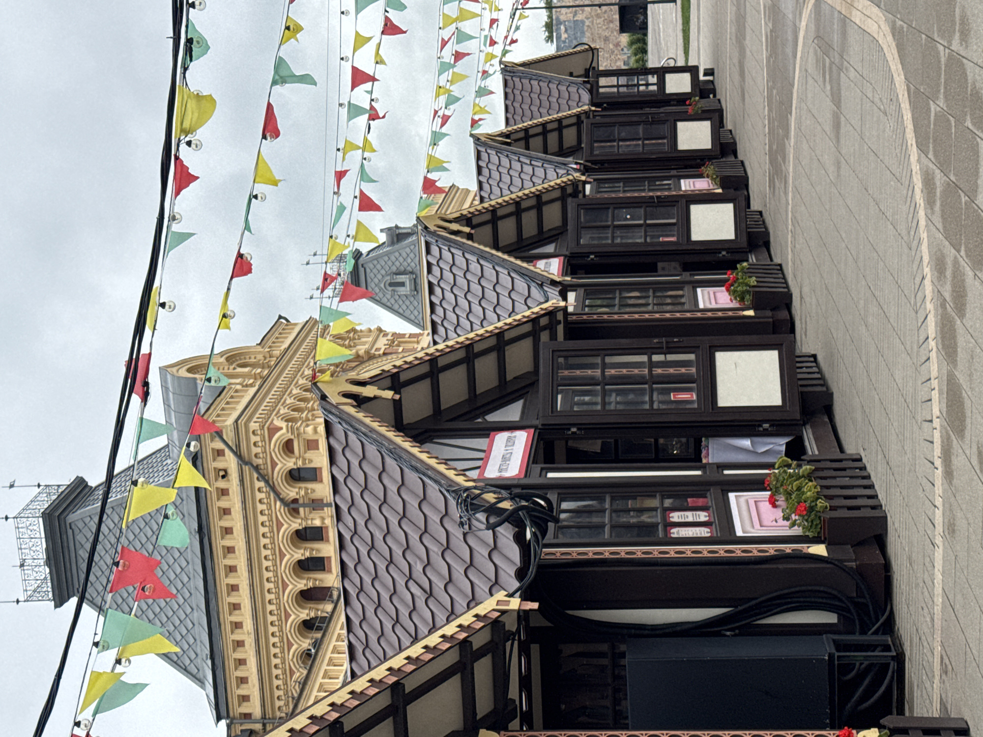 The image shows a row of small, dark brown wooden market stalls with brown tiled roofs, each featuring open windows and doors, situated on a paved outdoor area. In the background, a large, ornate building with a yellow facade and a dark grey roof with turrets stands prominently. Strings of colorful triangular flags in red, yellow, and green are strung across the scene, along with strings of small light bulbs. The stalls are lined up in a row, receding into the distance. Some stalls have flower boxes with red flowers and green foliage in front of them. The ground is paved with grey rectangular tiles, with some areas appearing damp. The sky is overcast and grey. This scene is located at the Nizhny Novgorod Kremlin in Nizhny Novgorod, Russia. There are no people visible in the image.