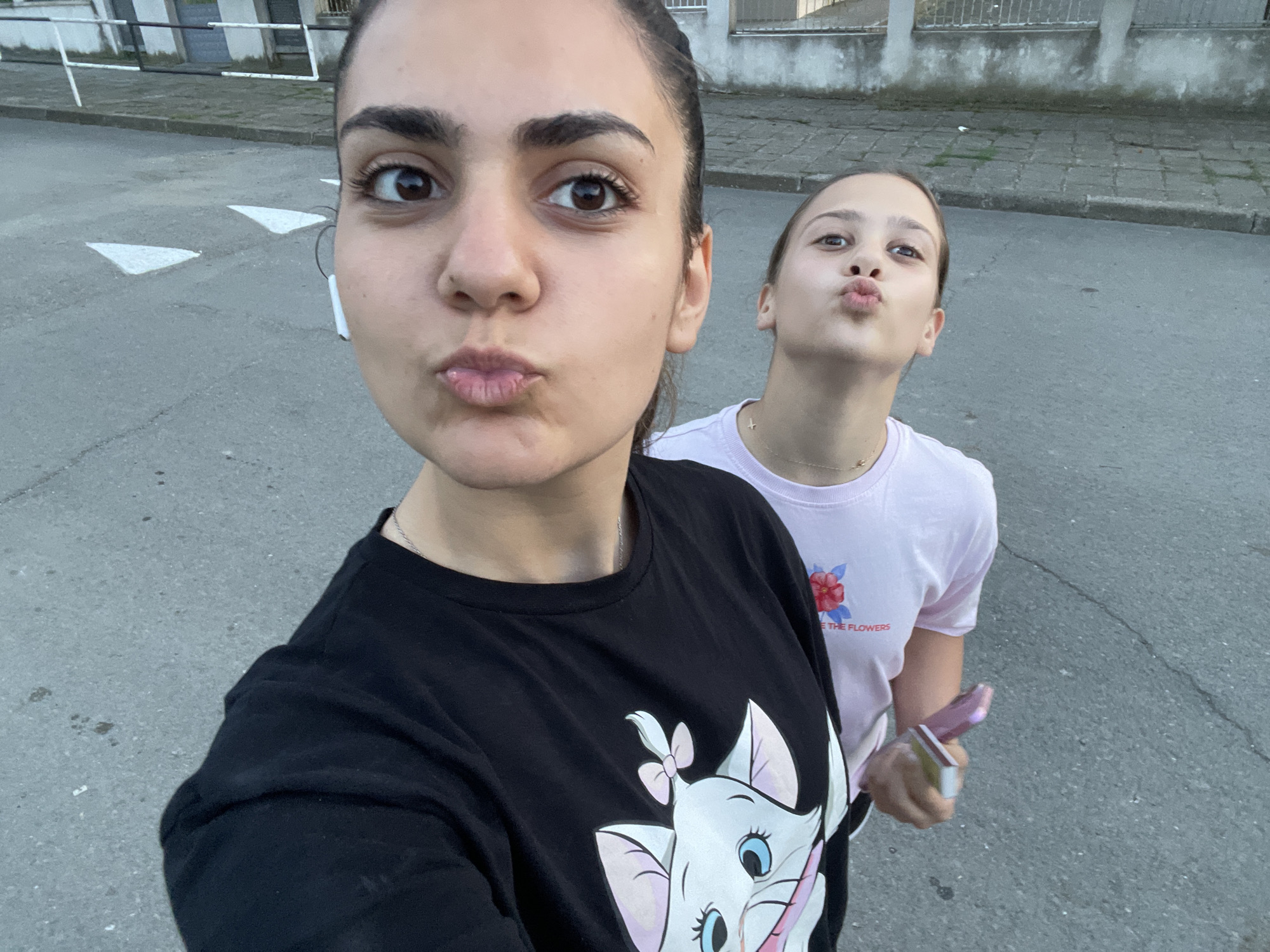 The scene is captured outdoors on an asphalt surface in Debelt, Bulgaria. Two females are posing for a close-up selfie, both pouting their lips towards the camera.
The primary subject, a young woman likely in her early twenties, occupies the foreground. She has dark brown hair pulled back and brown eyes. A white wireless earbud is visible in her right ear. She wears a black short-sleeved t-shirt featuring a large graphic of the white cartoon cat "Marie" from Disney's Aristocats on the chest. A thin silver chain necklace is visible around her neck.
Partially visible behind and to the right is a younger female, likely a teenager. She also pouts her lips towards the camera. She wears a light pink short-sleeved t-shirt adorned with a red floral graphic and partially legible text "E THE FLOWERS" on the left chest. A delicate gold-colored chain necklace with a small charm is around her neck. Her right hand is visible, holding a pink smartphone-like device and a smaller, rectangular silver object, possibly a compact electronic accessory.
The ground is dark grey asphalt, showing some minor cracks and debris. To the left of the primary subject, two white triangular road markings are painted on the asphalt. In the background, a light-colored concrete wall or fence with horizontal bars is visible, separating the street from what appears to be a building or residential area. The lighting suggests an outdoor setting during soft daylight hours.