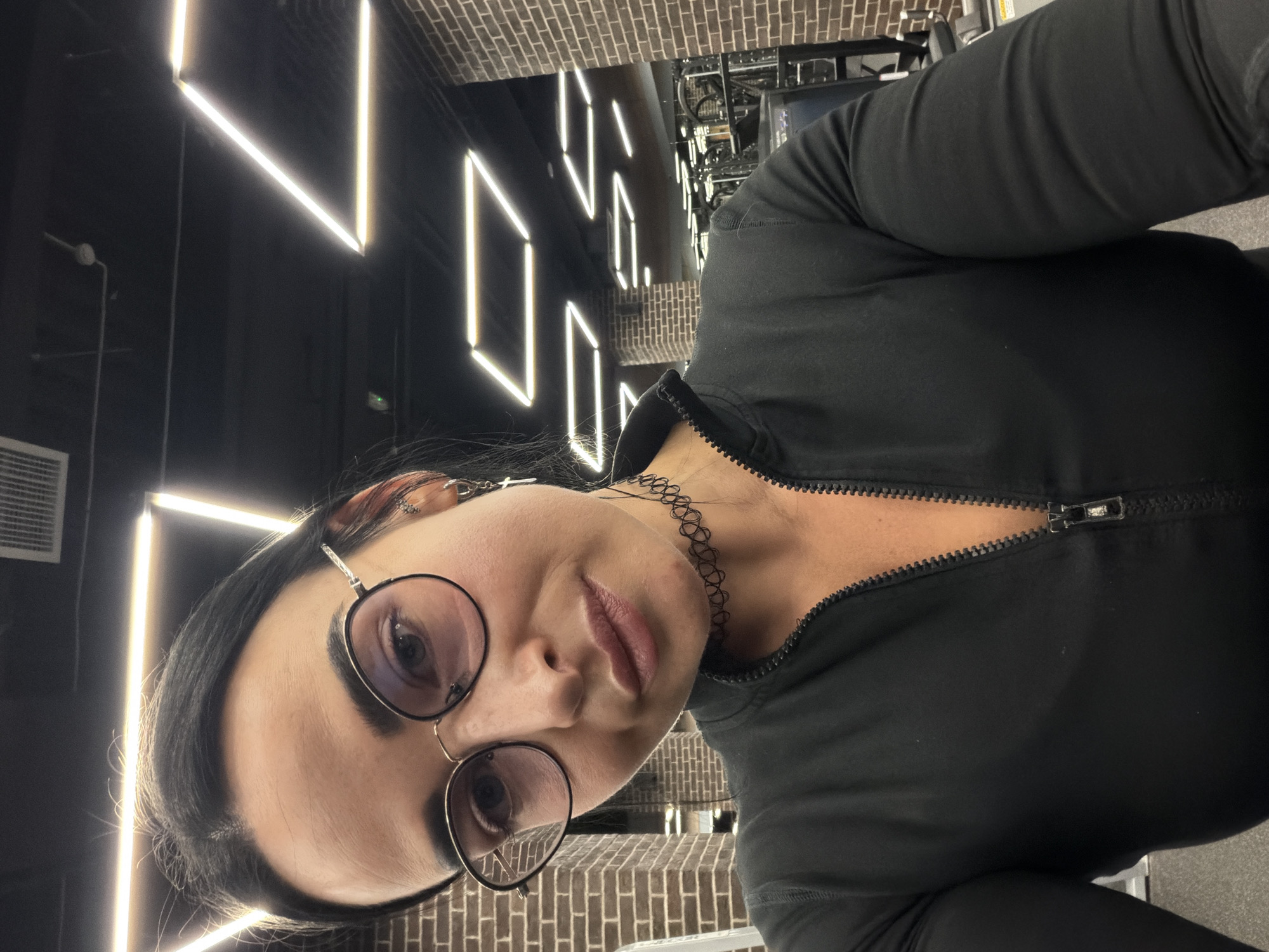 A woman with dark hair and glasses is captured in a close-up selfie within a modern fitness center in Saint Petersburg, Russia. She gazes directly into the camera with a neutral expression, her lips slightly parted, and her dark hair parted on the viewer's left side. She wears round, thin-framed metal glasses and a black long-sleeved athletic top with a prominent zipper running down the front, partially unzipped. Accessories include a black, stretchy tattoo choker necklace around her neck and multiple earrings on her visible right ear, specifically a hoop earring with a dangling white cross charm. The background features exposed reddish-brown brick walls on the left and right, creating an industrial aesthetic. The dark ceiling is illuminated by an array of bright, linear LED light fixtures arranged in various geometric shapes, including squares and parallelograms. An air vent is visible in the upper left corner of the ceiling. In the distant background, indistinct gym equipment is visible, confirming the setting as a fitness facility. The scene is well-lit by the overhead artificial lighting.
