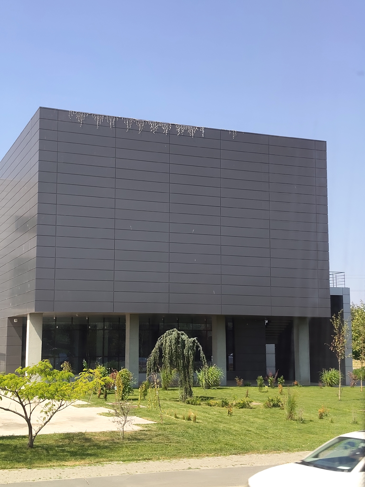 A modern, gray, rectangular building with large windows on the ground floor stands under a clear blue sky. The building's facade is composed of horizontal panels, and at the very top, there is a string of white, icicle-like decorations. In front of the building, a well-maintained lawn is dotted with various trees and shrubs, some of which have weeping branches. A concrete walkway is visible in the foreground, and a portion of a white car can be seen on the right side of the image. The scene appears to be an exterior shot of a business or institutional building in a city, possibly during an off-season given the decorations.